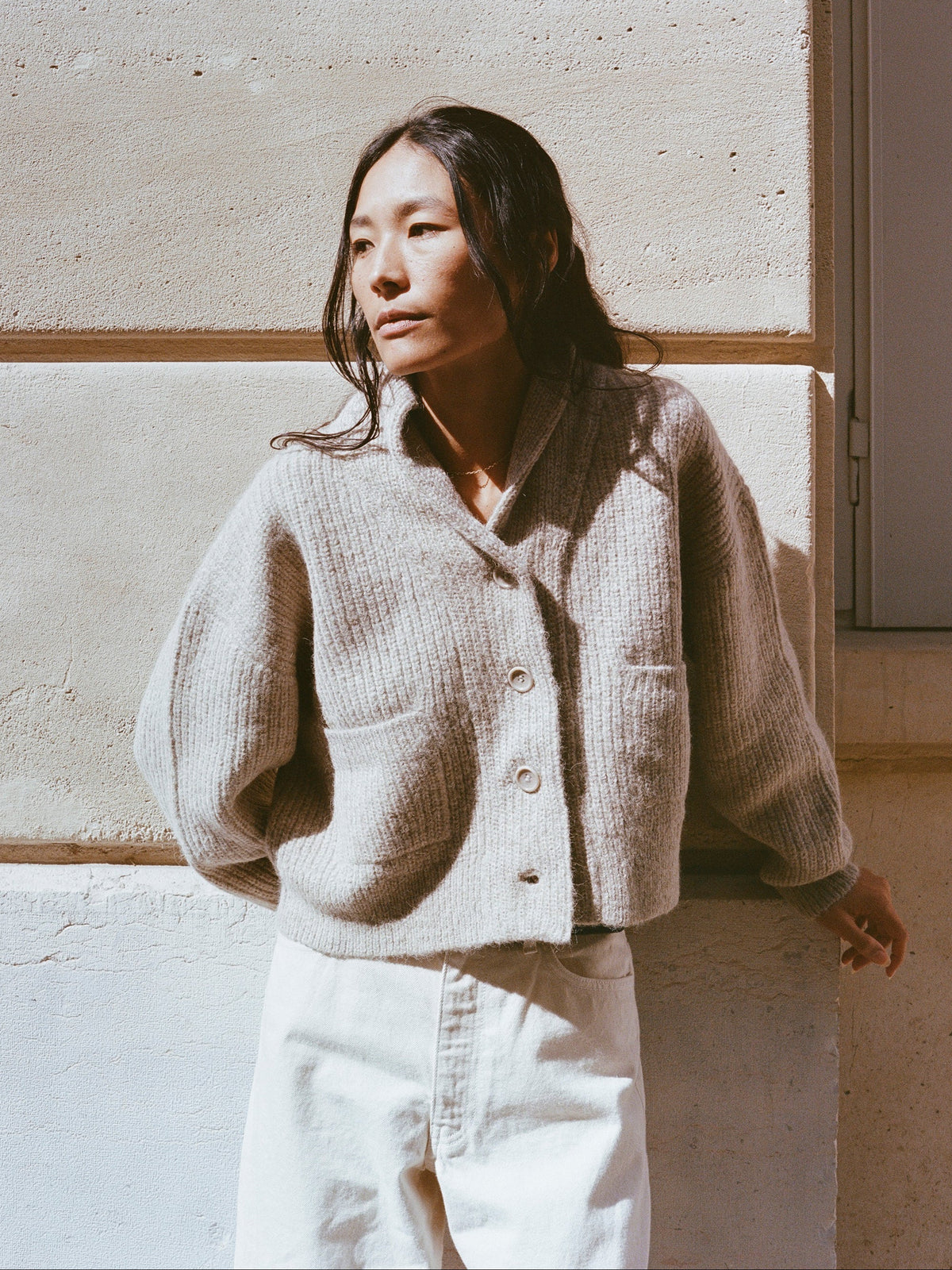 Woman wearing a beige knitted cardigan and white pants against a light-colored wall.