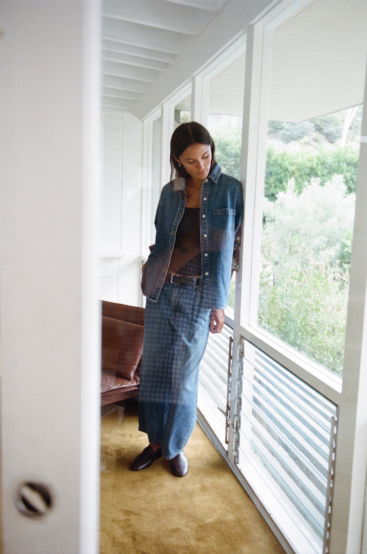 A woman stands in a bright interior space with large windows, wearing a long blue denim dress layered over a brown top and dark shoes. The photo is taken through a doorway, with white walls, a tan carpet, and greenery visible outside.