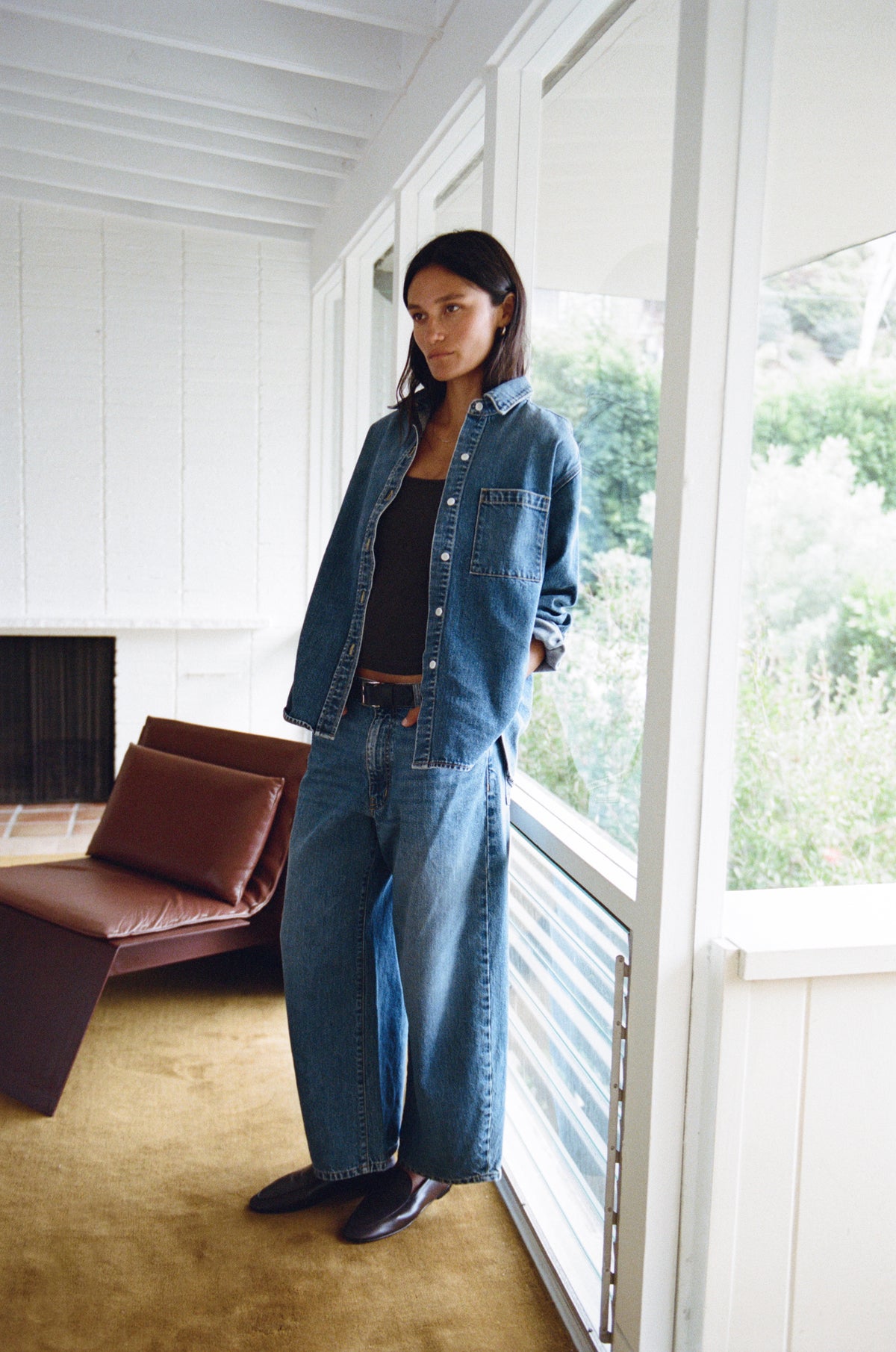A woman stands on a sunlit porch wearing a blue denim button-up shirt and matching wide-leg jeans over a black top, with a brown leather chair and large windows in the background.