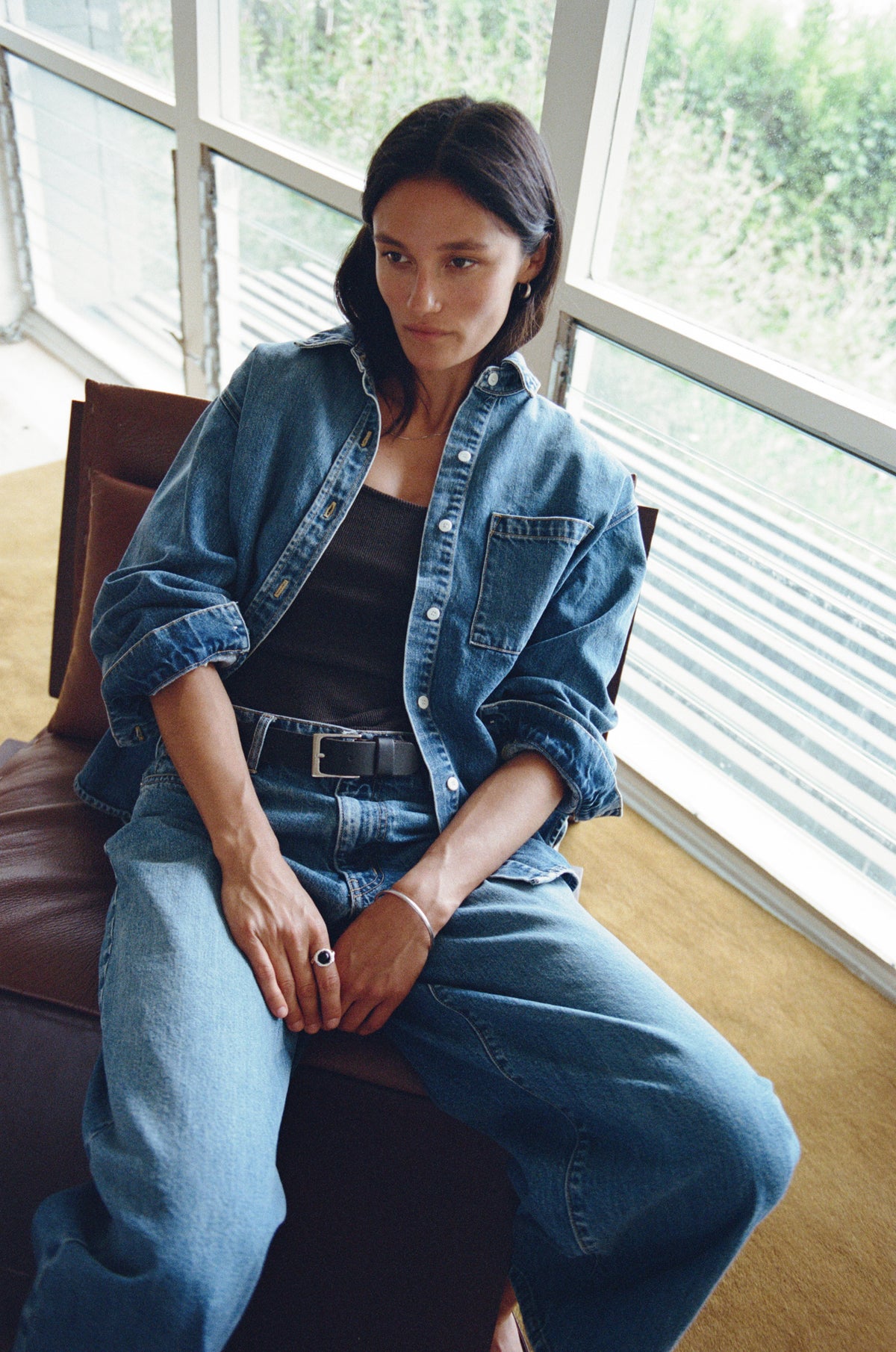 A woman sits on a brown leather chair in a bright room with large windows, wearing a blue denim shirt and matching wide-leg jeans over a black top.