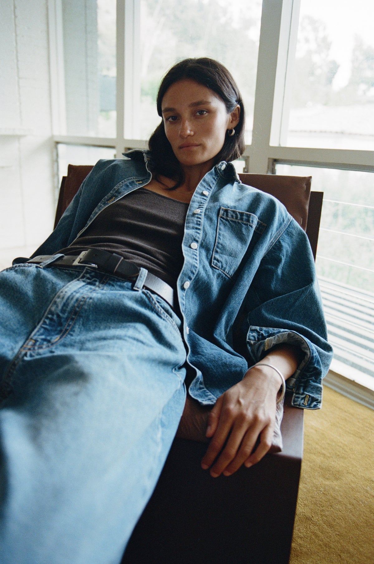 A woman reclining in a chair, wearing a blue denim shirt over a black top and blue jeans, in a brightly lit indoor setting with large windows.