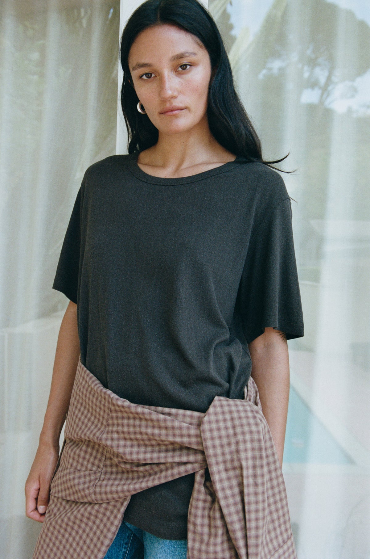 A woman wearing an oversized dark charcoal gray t-shirt and a brown and beige gingham shirt tied around her waist, standing in front of a soft-focus background with sheer curtains.