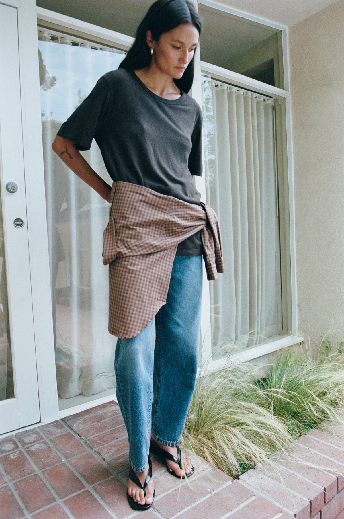 A woman stands on a brick porch wearing a dark charcoal gray t-shirt, blue jeans, and black flip-flops, with a brown and beige gingham shirt tied around her waist.
