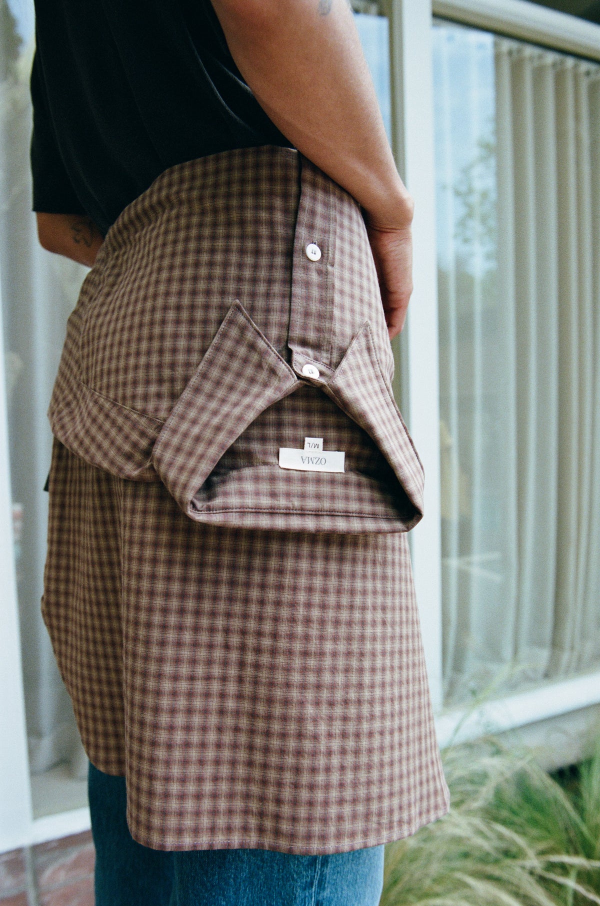 A person wearing a brown and beige gingham overshirt over a black shirt and blue jeans, standing in front of a window.