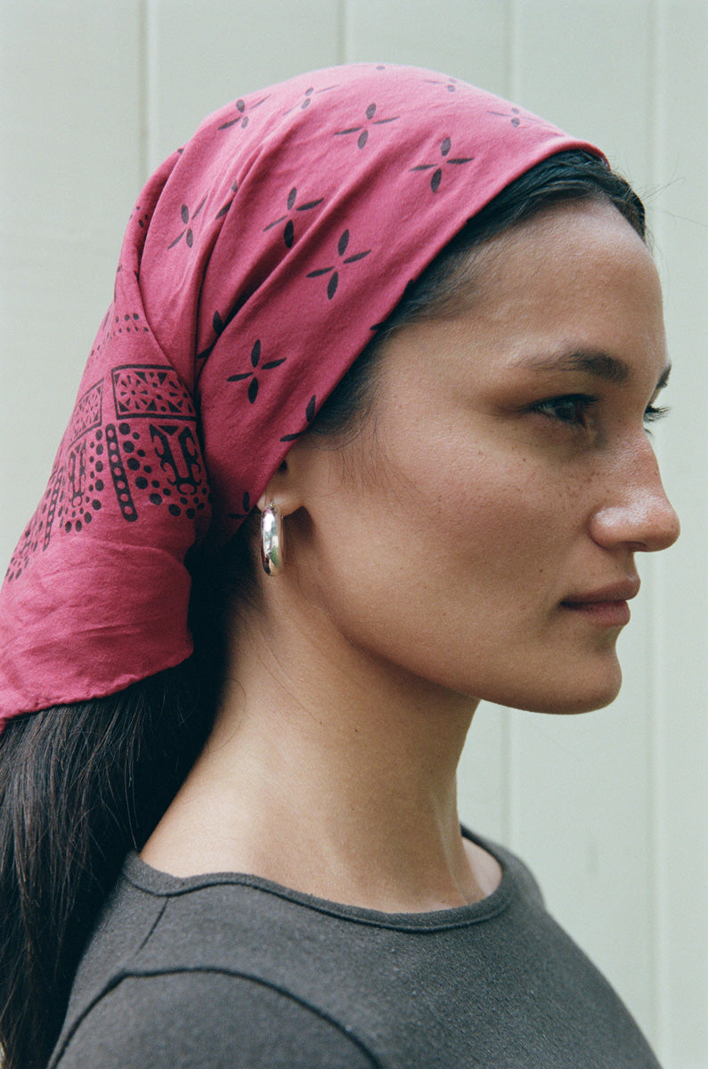 A woman in profile wearing a pink bandana headscarf with black geometric patterns, paired with a dark gray shirt and silver hoop earrings, set against a light gray paneled wall.