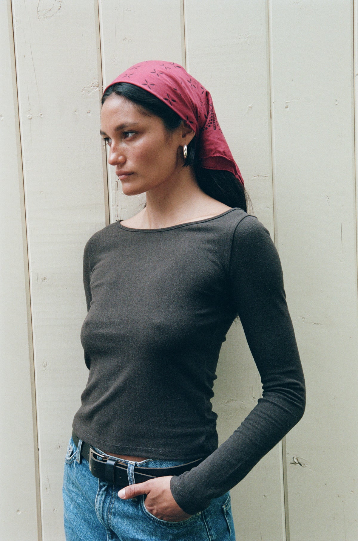 A woman wearing a dark charcoal gray long-sleeve boatneck top, blue jeans, and a dusty red patterned headscarf, standing against a light beige wooden wall.