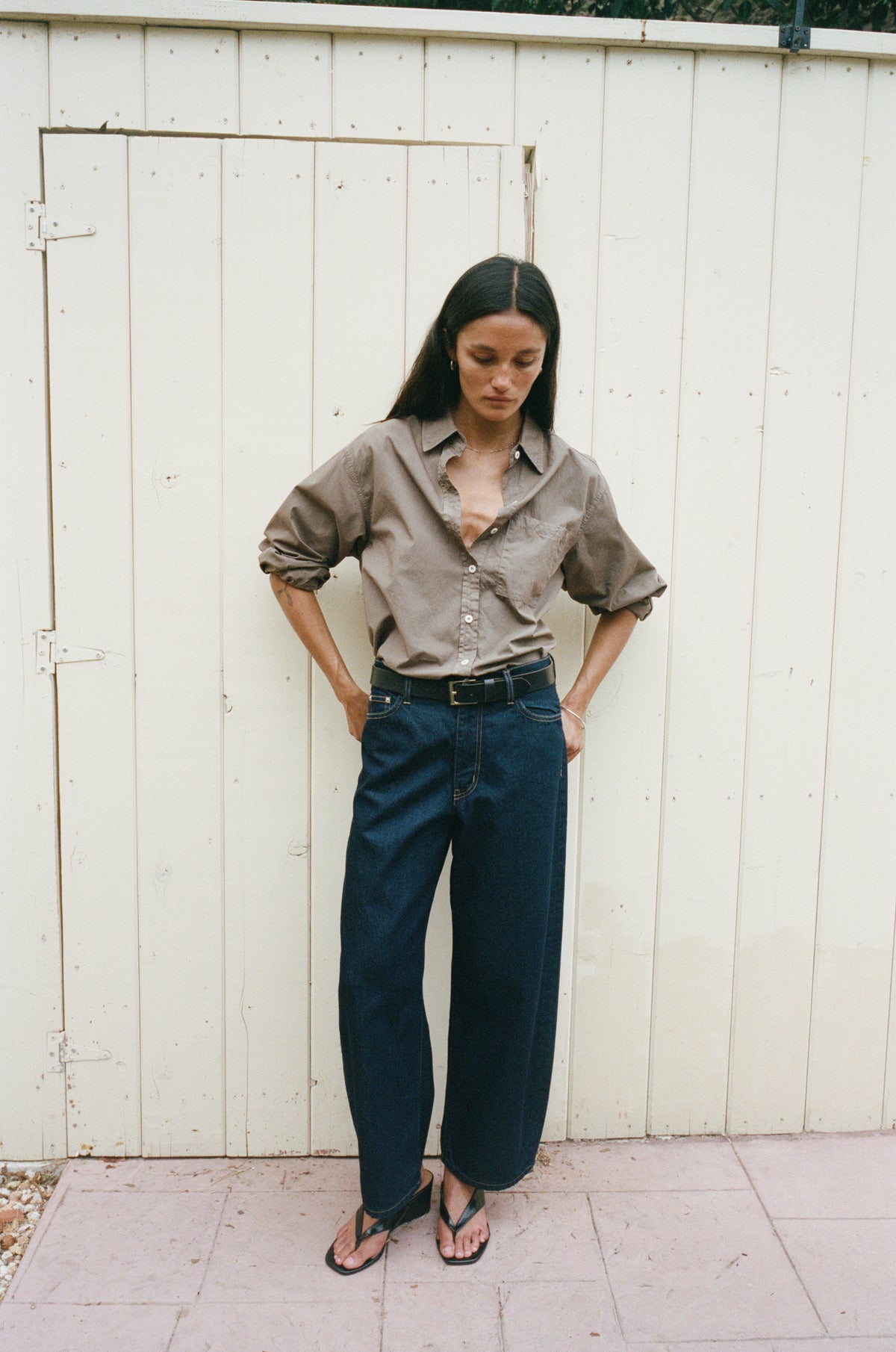 A woman stands against a cream-colored wooden fence wearing a brown button-down shirt, dark blue wide-leg jeans, and black sandals.