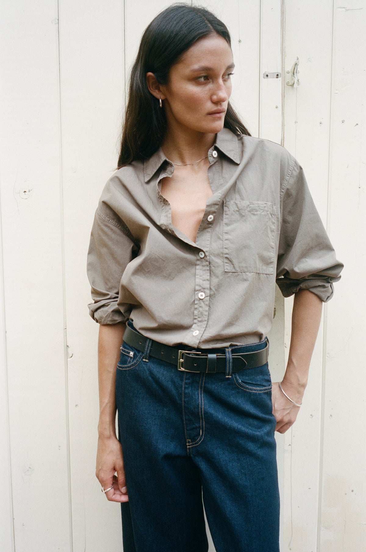 A woman wearing a light brown button-down shirt with rolled-up sleeves and dark blue jeans, standing against a white wooden wall.