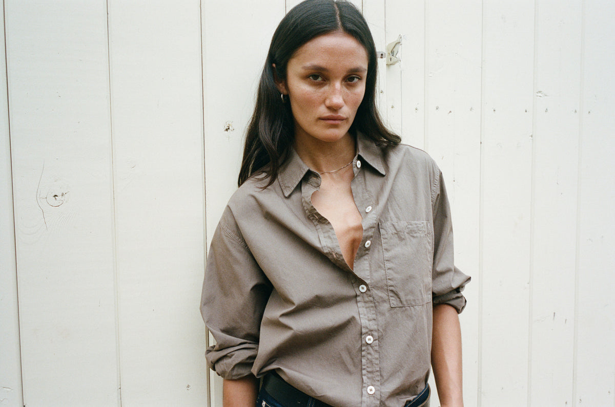 A woman with long dark hair stands against a white wooden wall wearing a muted taupe-brown button-down shirt with rolled-up sleeves and dark jeans.