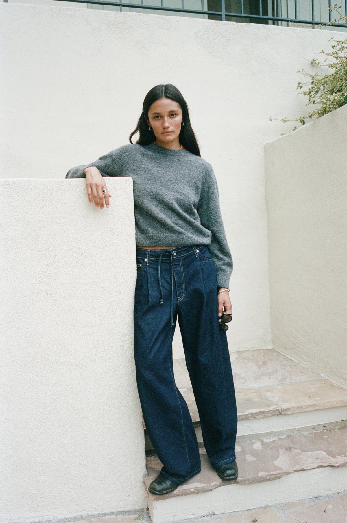 A woman stands on outdoor stone steps against a white stucco wall, wearing a gray long-sleeve knit sweater and dark wide-leg jeans.