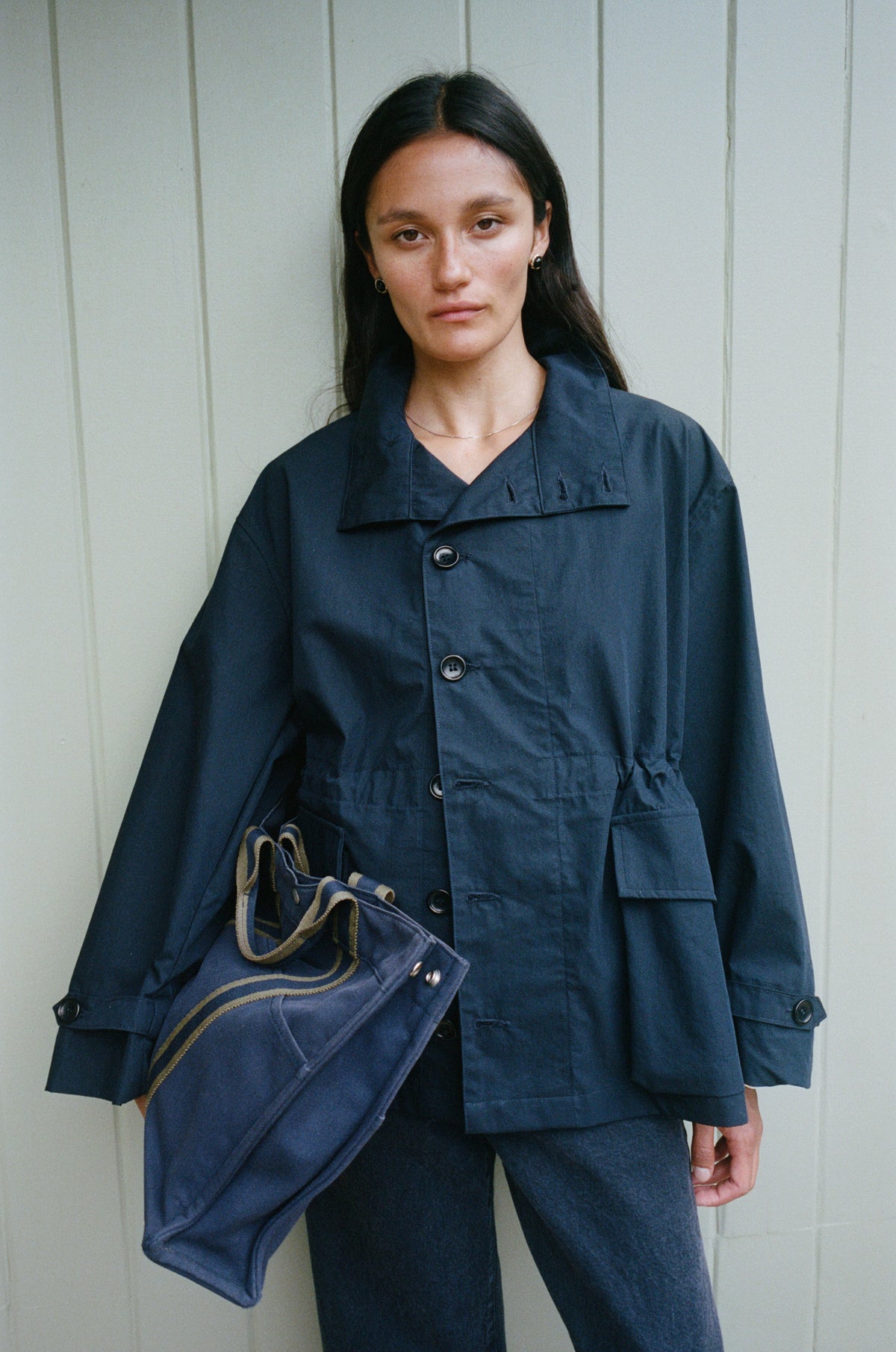 A woman wearing a dark navy blue utility jacket and matching pants, holding a matching navy blue tote bag, standing against a light gray paneled wall.