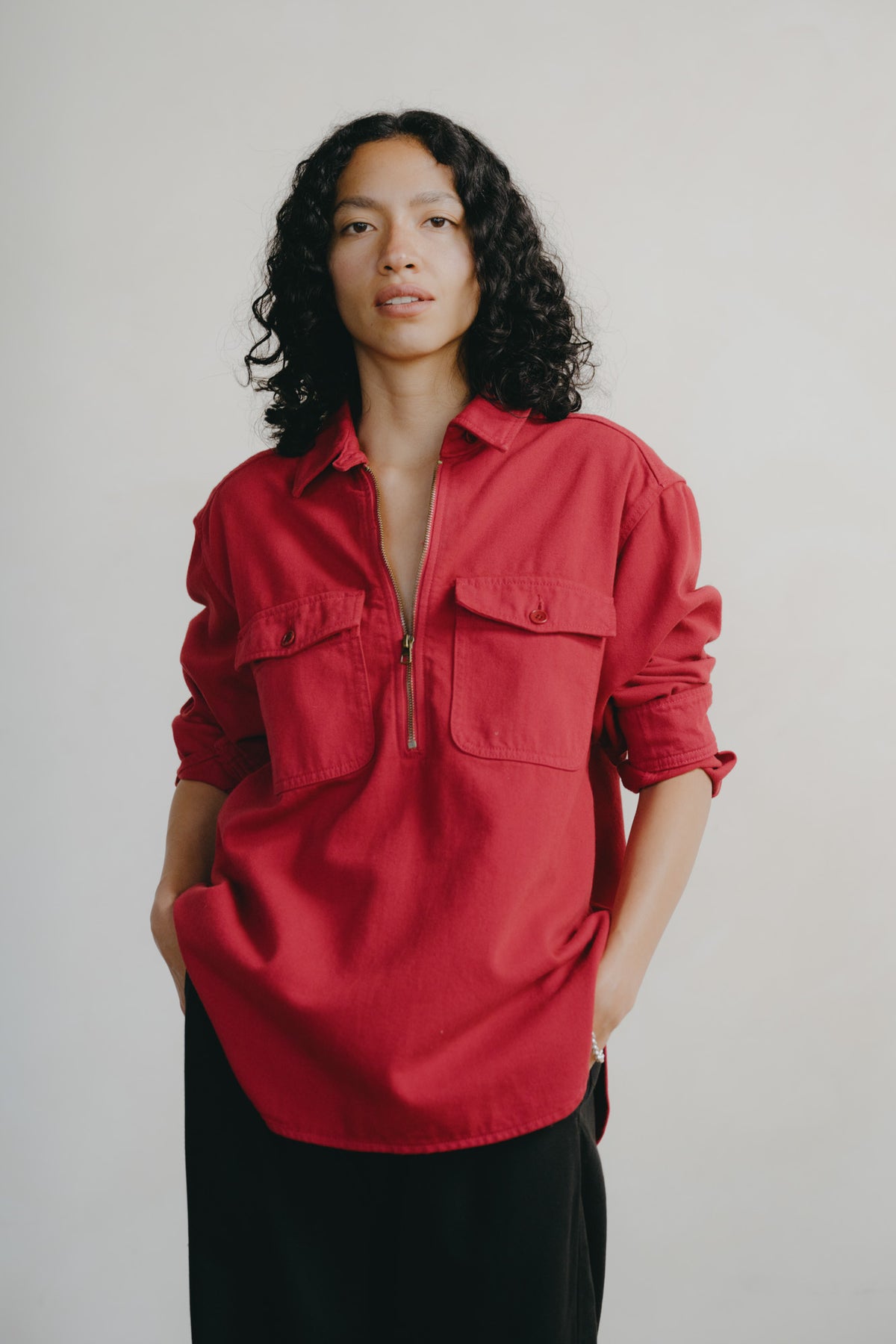 A model with curly black hair wearing a red button-down shirt with a half-zip front and black pants, standing against a plain light gray background.