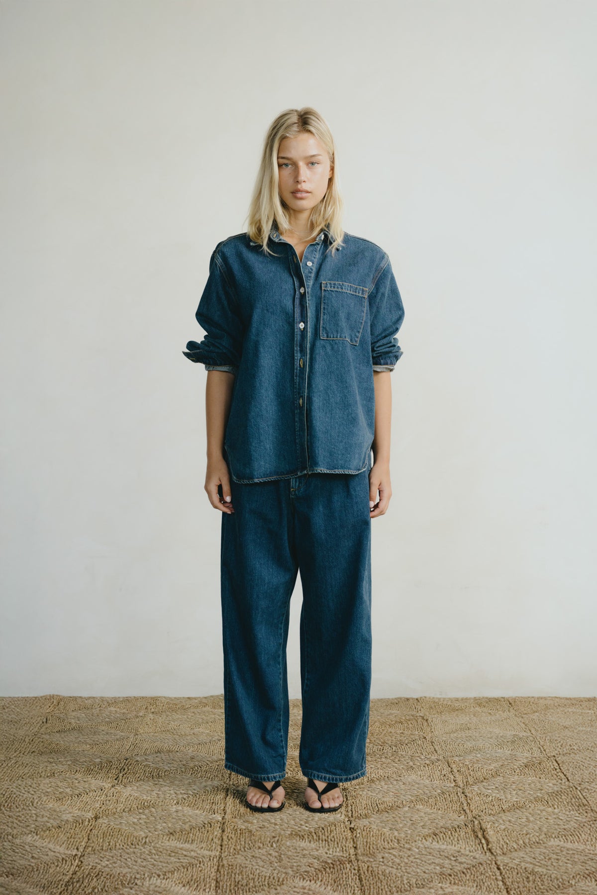 A model stands against a light off-white wall wearing a matching dark blue denim set consisting of a button-up shirt and wide-leg pants, paired with black sandals on a textured straw-colored floor.