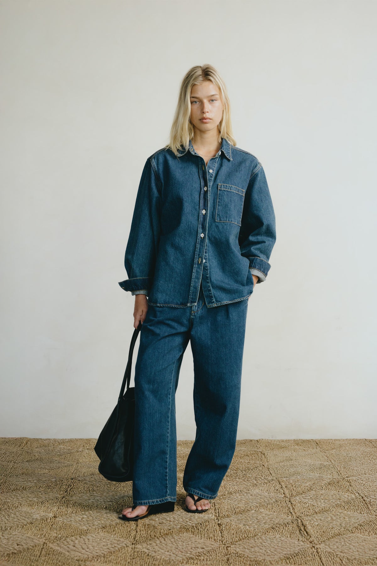 A woman stands in a studio wearing an oversized dark blue denim shirt and matching wide-leg denim pants, holding a black tote bag. The background is a clean off-white wall with a textured tan floor.