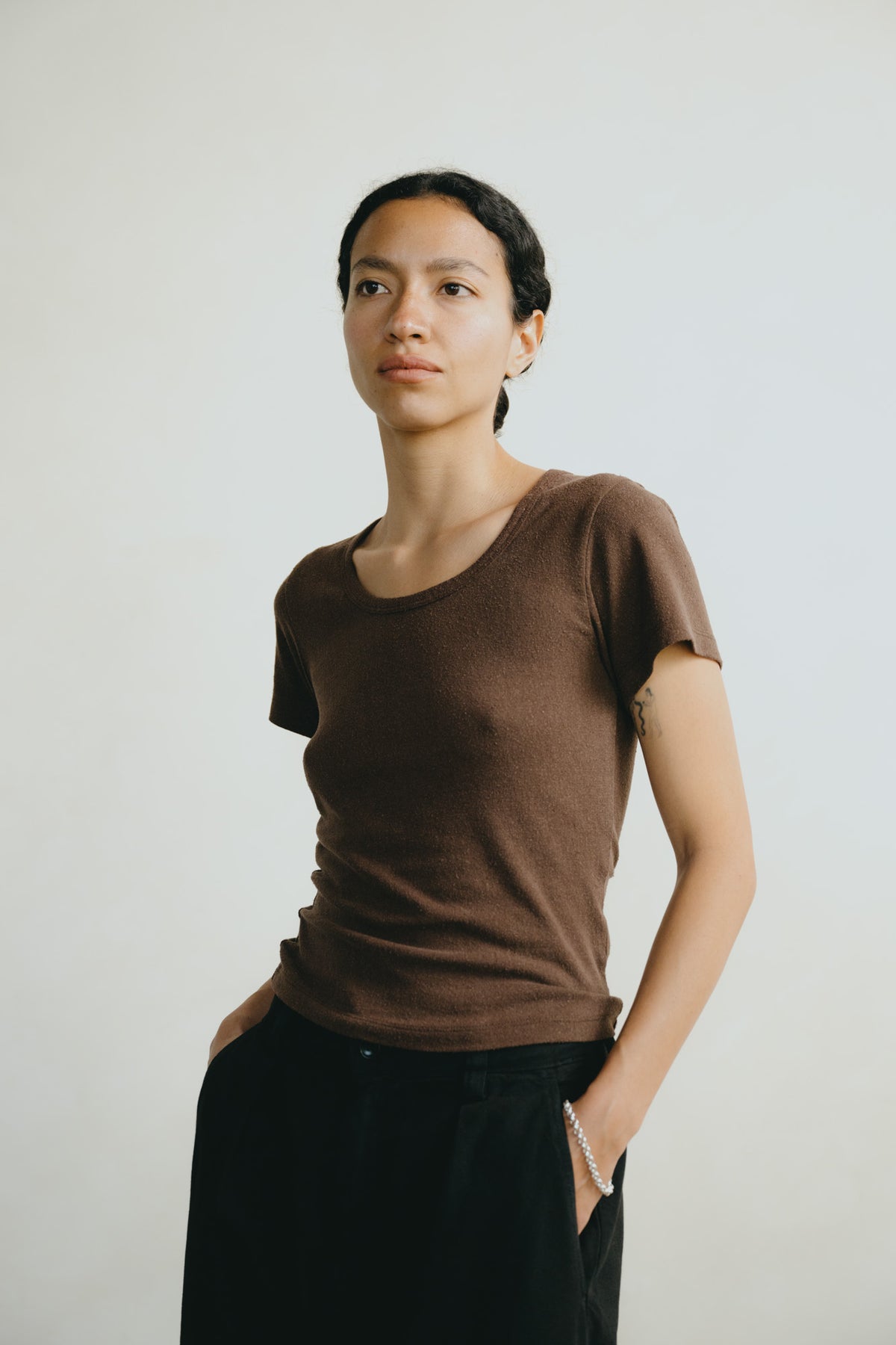 A woman wearing a brown short-sleeve crewneck t-shirt and black pants, standing against a plain light gray background.