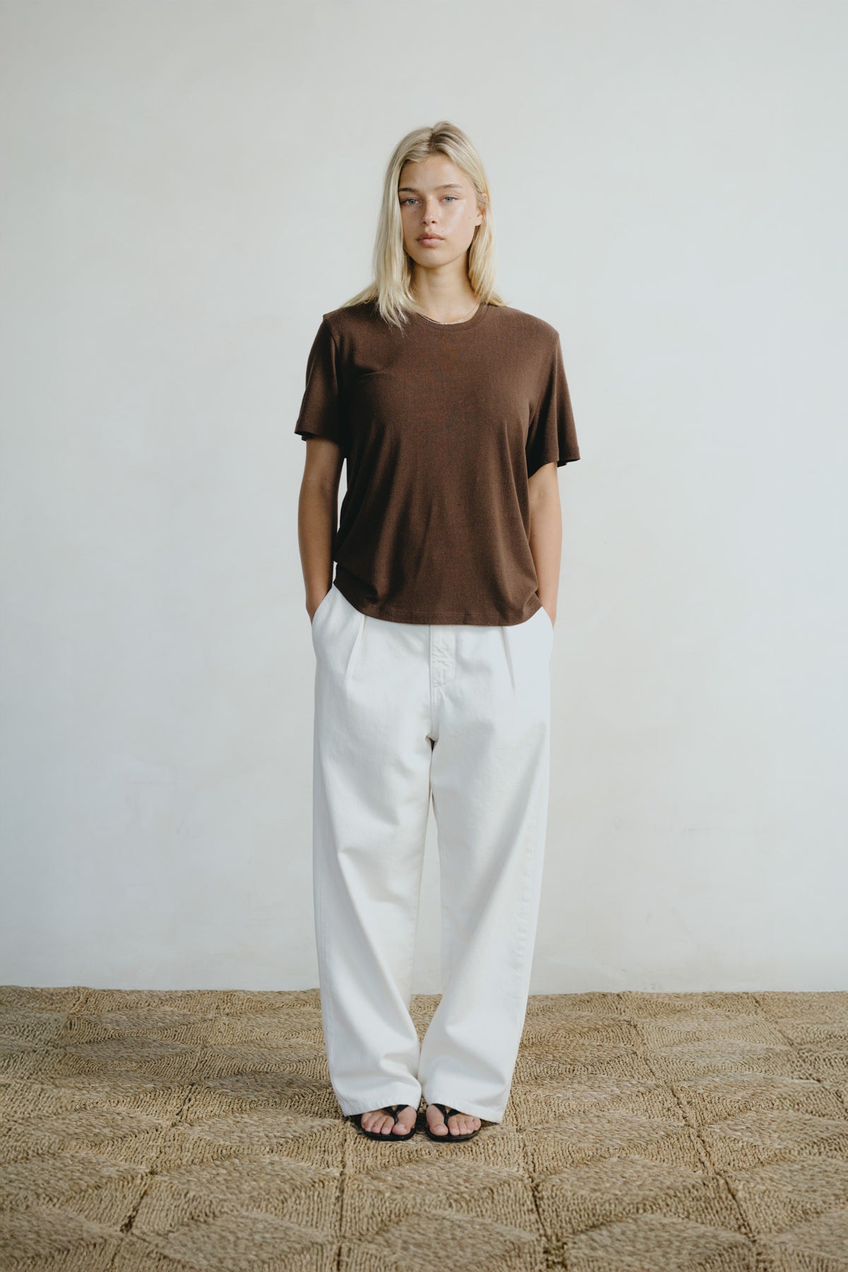 A woman stands against a plain off-white wall wearing a brown short-sleeve crewneck t-shirt and loose white wide-leg pants, with her hands in her pockets.