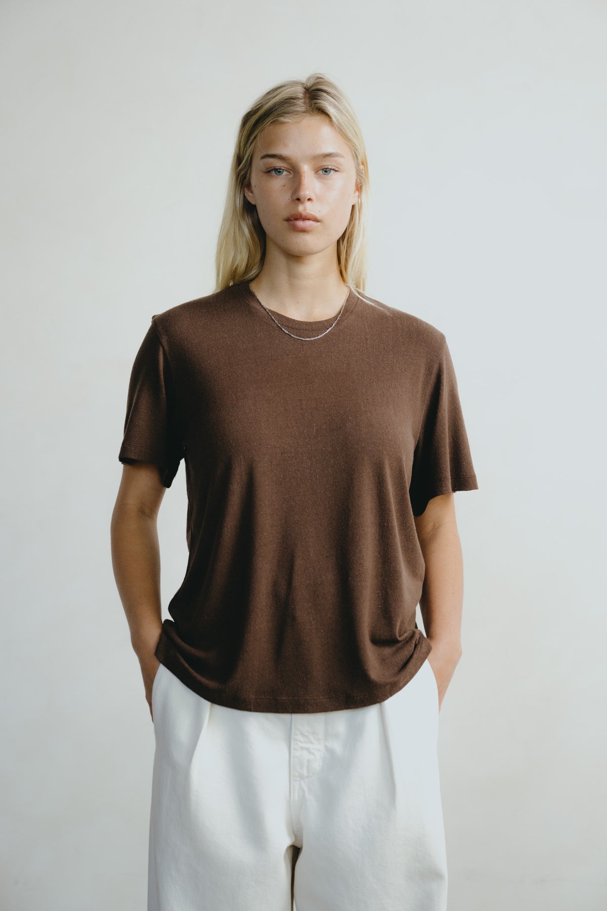 A woman modeling a brown short-sleeve crewneck t-shirt with white pants, standing against a light gray studio background.