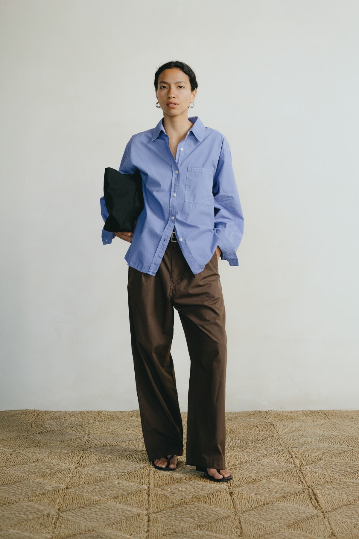 A model stands in a studio setting wearing a light blue button-down shirt and dark brown wide-leg trousers, holding a black clutch bag against a neutral off-white wall and textured beige floor.