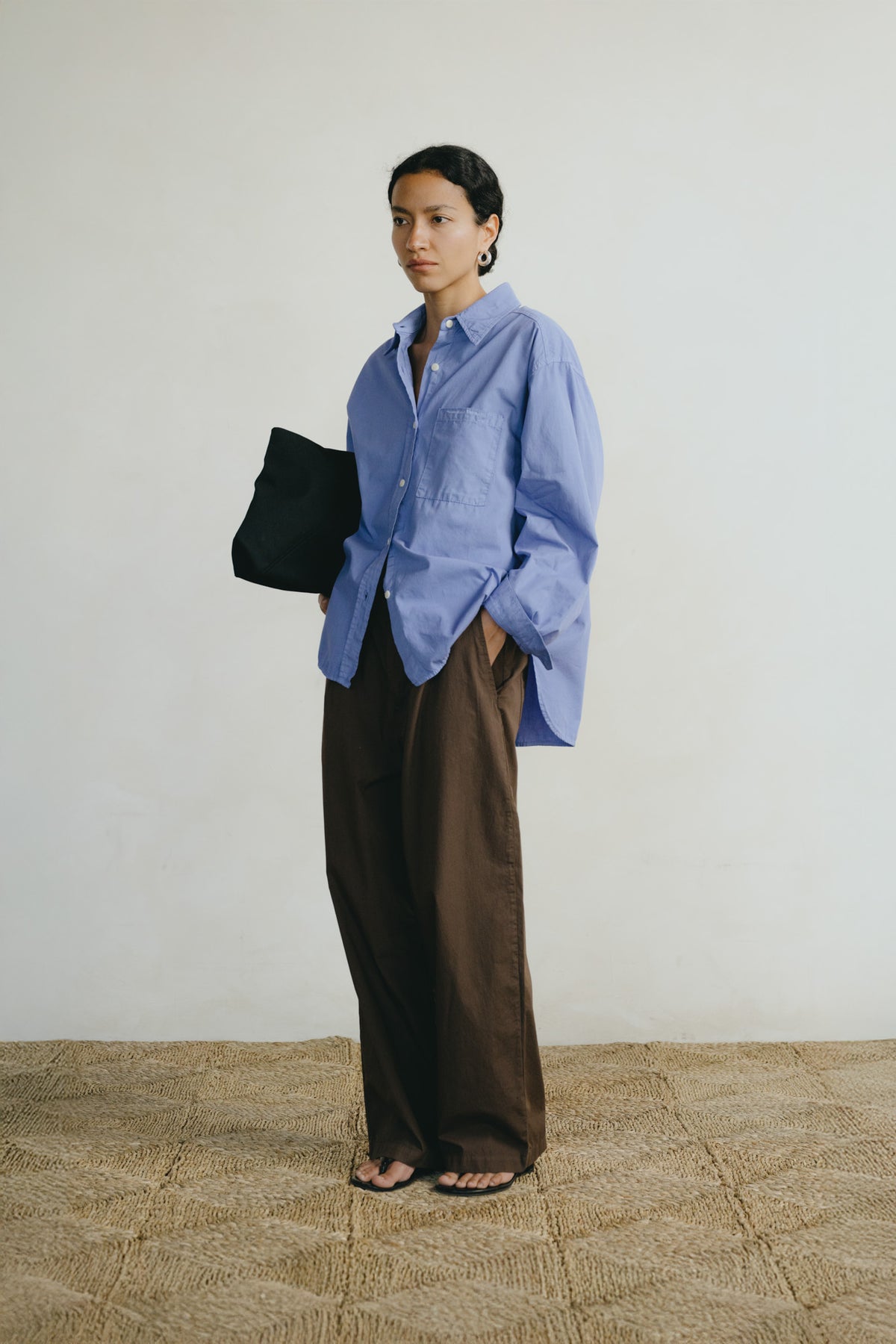 A model stands in a minimalist room wearing a light blue button-down shirt and dark brown wide-leg trousers, holding a black tote bag.