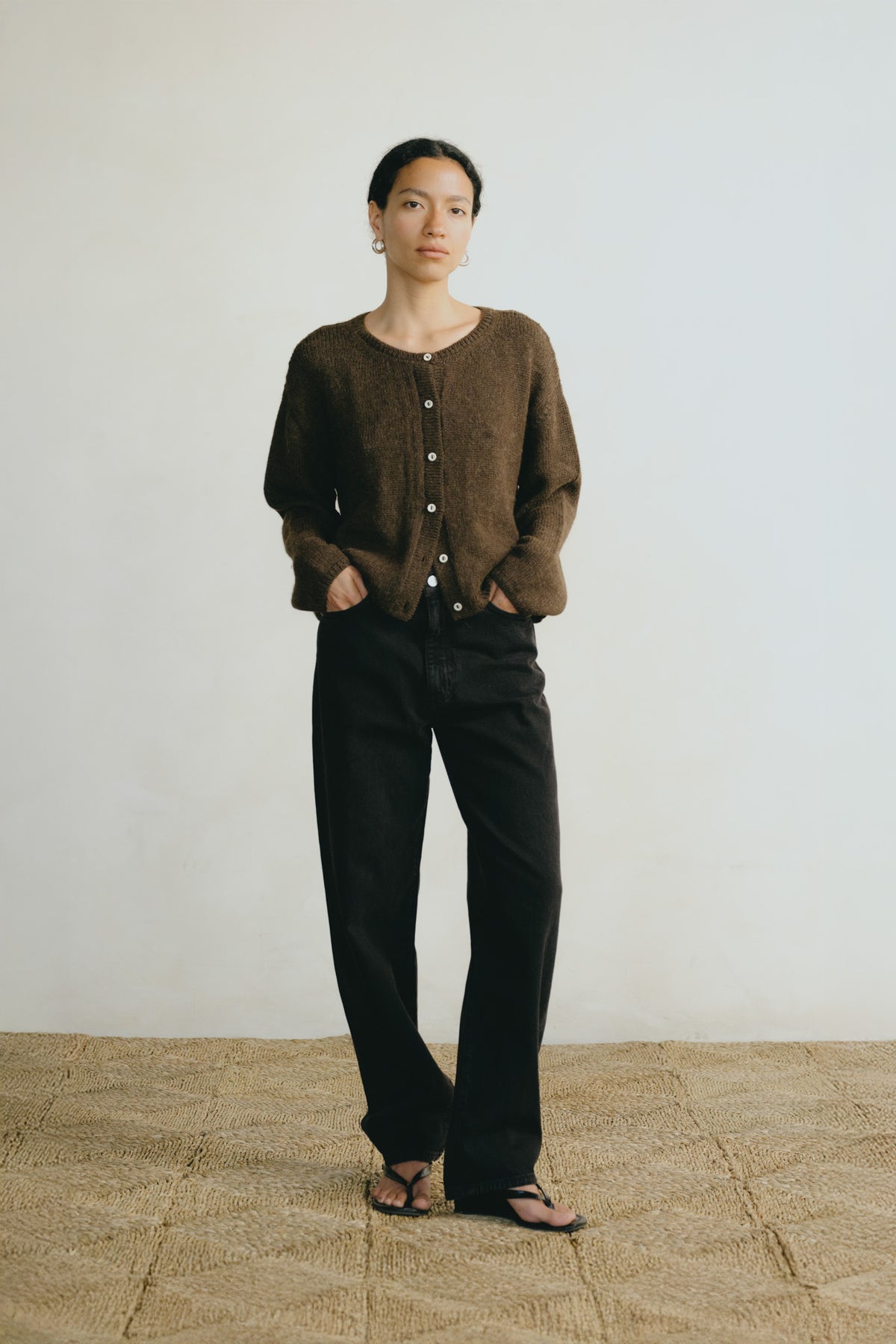 A model stands against a plain off-white wall wearing a brown fuzzy button-front cardigan, black wide-leg pants, and black sandals on a textured beige floor.