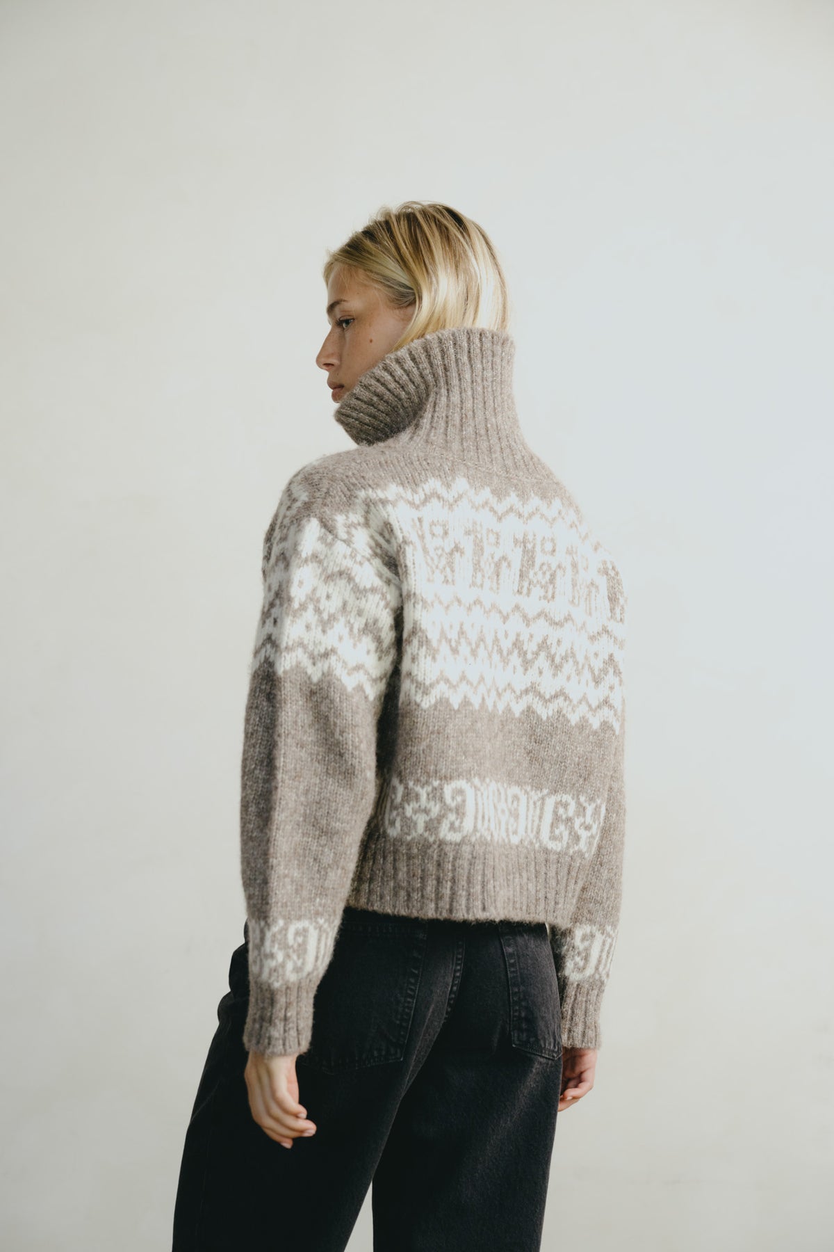 A woman shown from the back wearing a cropped turtleneck sweater with a cream Fair Isle pattern on a taupe-brown base, paired with black jeans, against a plain light gray studio background.