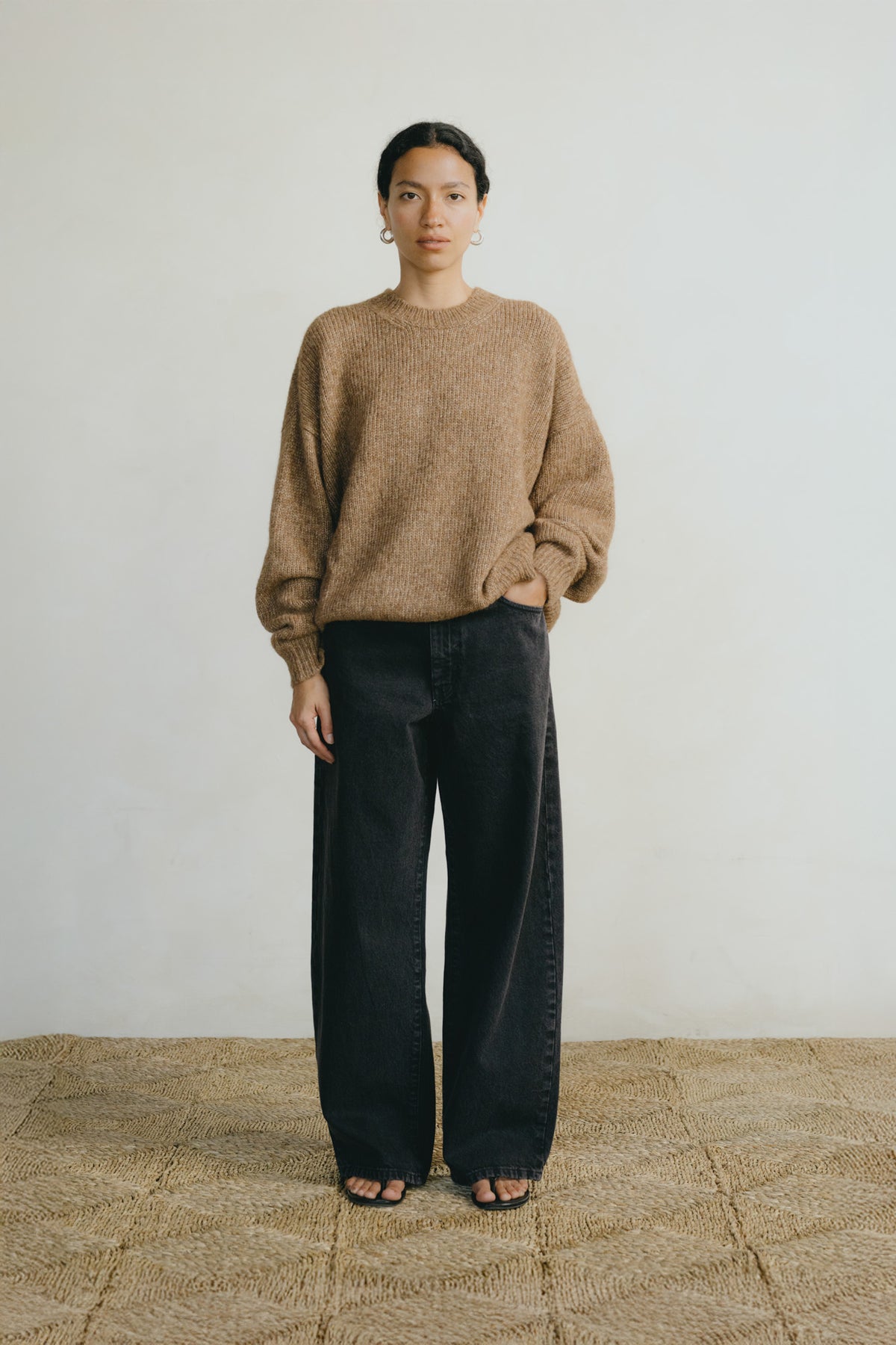 A woman stands against a plain off-white wall wearing a loose, fuzzy taupe-brown knit sweater and wide-leg black jeans, with sandals on a woven beige floor.