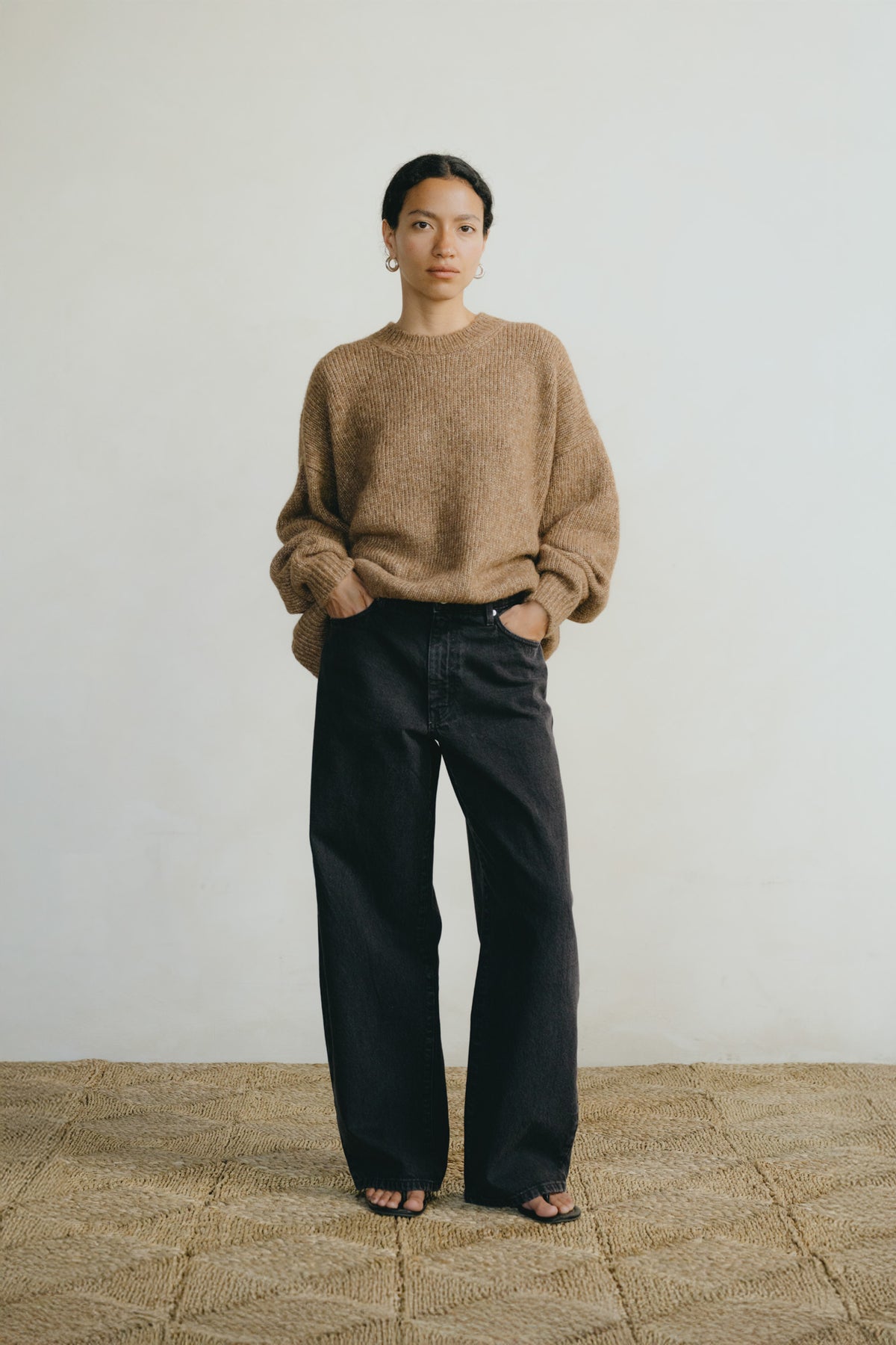 A woman stands against a plain off-white wall wearing a loose, fuzzy taupe-brown sweater and wide-leg black jeans, with sandals on a woven straw floor.