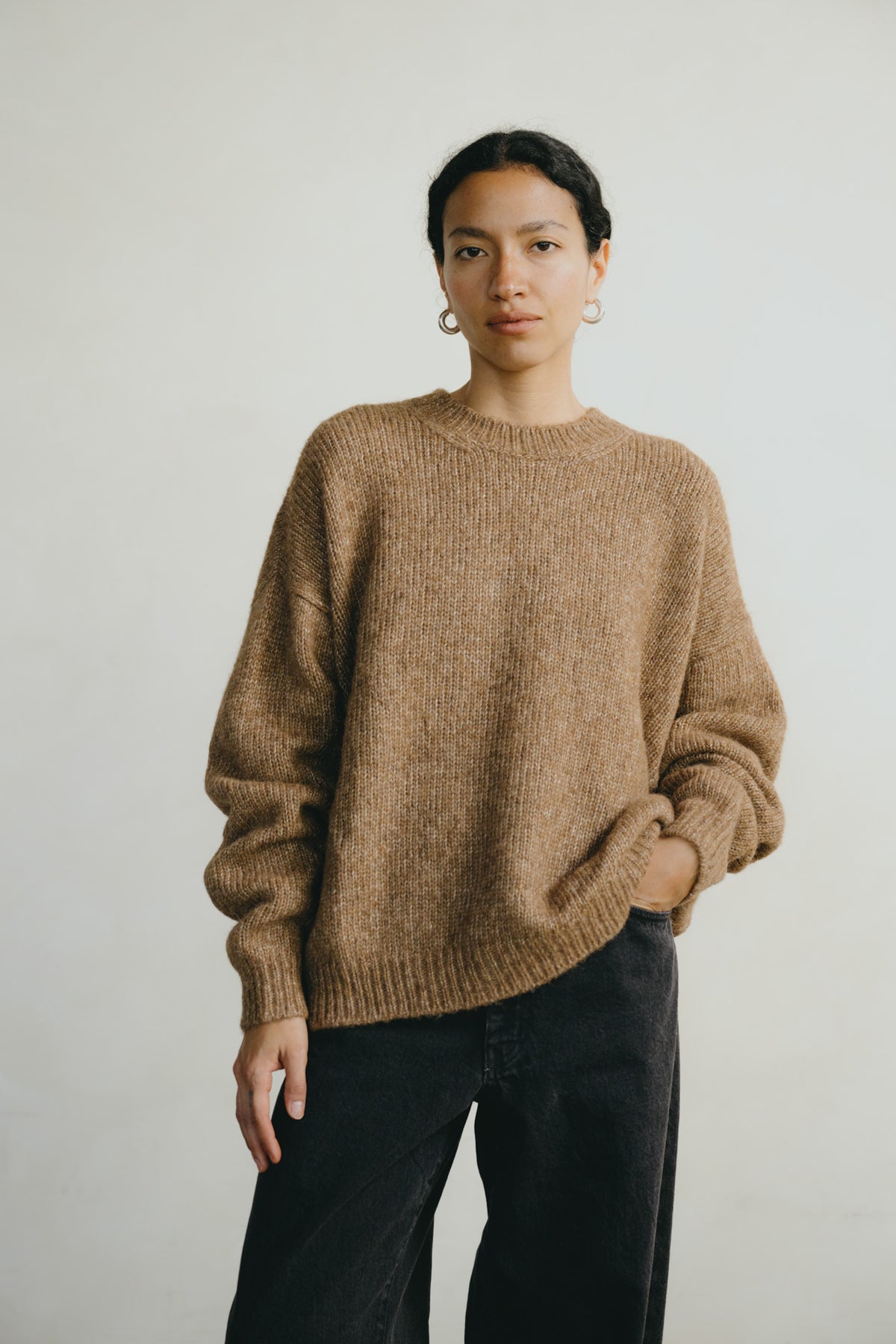 A model wearing a textured, oversized brown knit sweater and dark charcoal pants, standing against a plain light gray background.