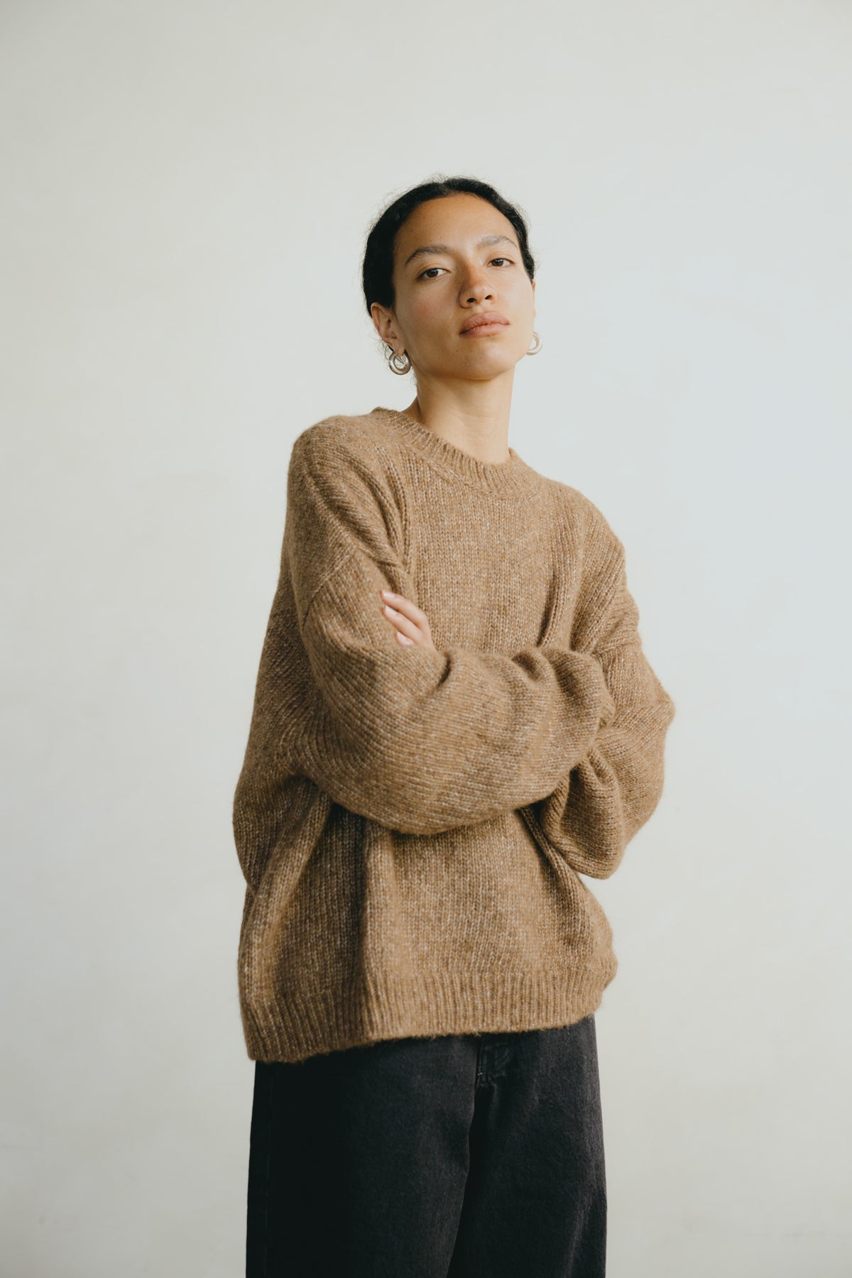 A person wearing an oversized, chunky knit crewneck sweater in a warm camel brown color, paired with dark pants, standing against a plain light gray background.