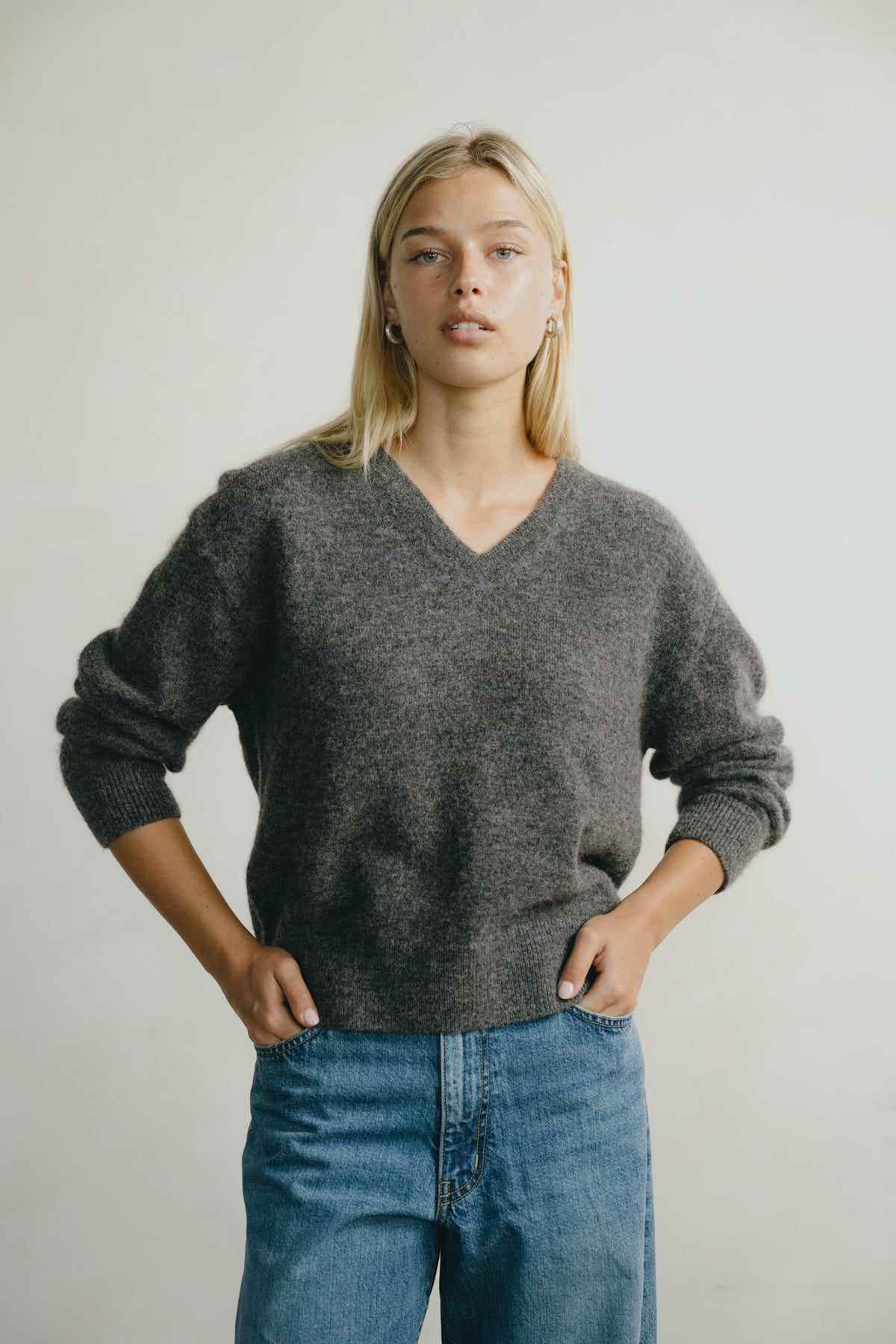 A woman wearing a dark gray v-neck knit sweater and blue jeans, standing against a plain light gray background.