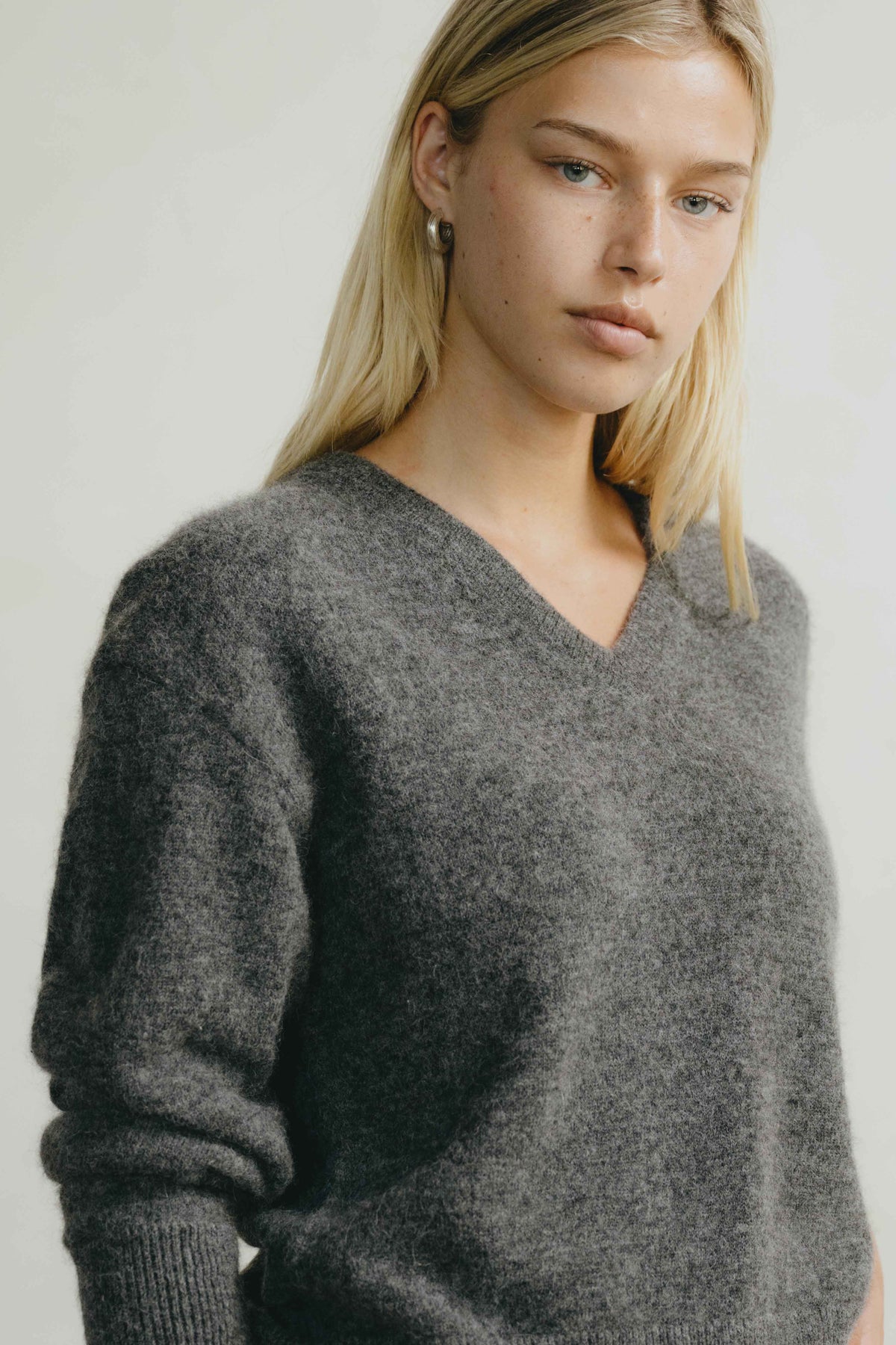 A blonde woman wearing a fuzzy dark gray V-neck knit sweater, photographed against a plain light background.