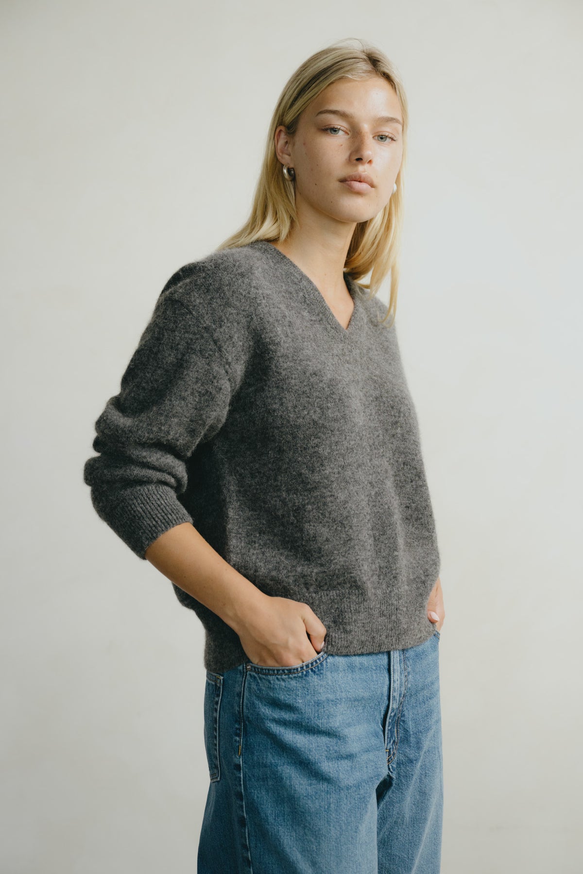 A woman wearing a dark gray fuzzy V-neck sweater and blue jeans, standing against a plain light gray background.
