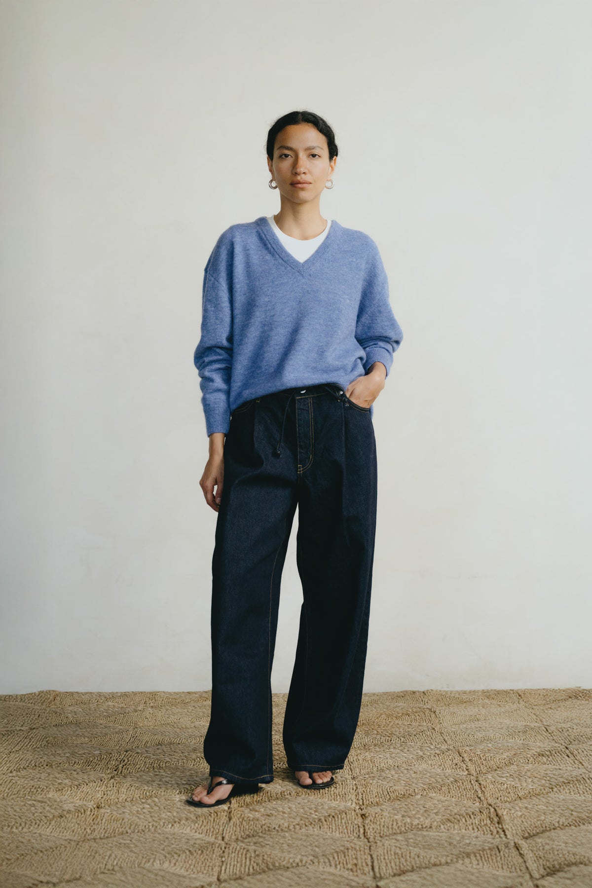 A woman stands in a minimalist studio setting wearing a light blue v-neck knit sweater over a white undershirt, paired with dark wide-leg trousers and black sandals.