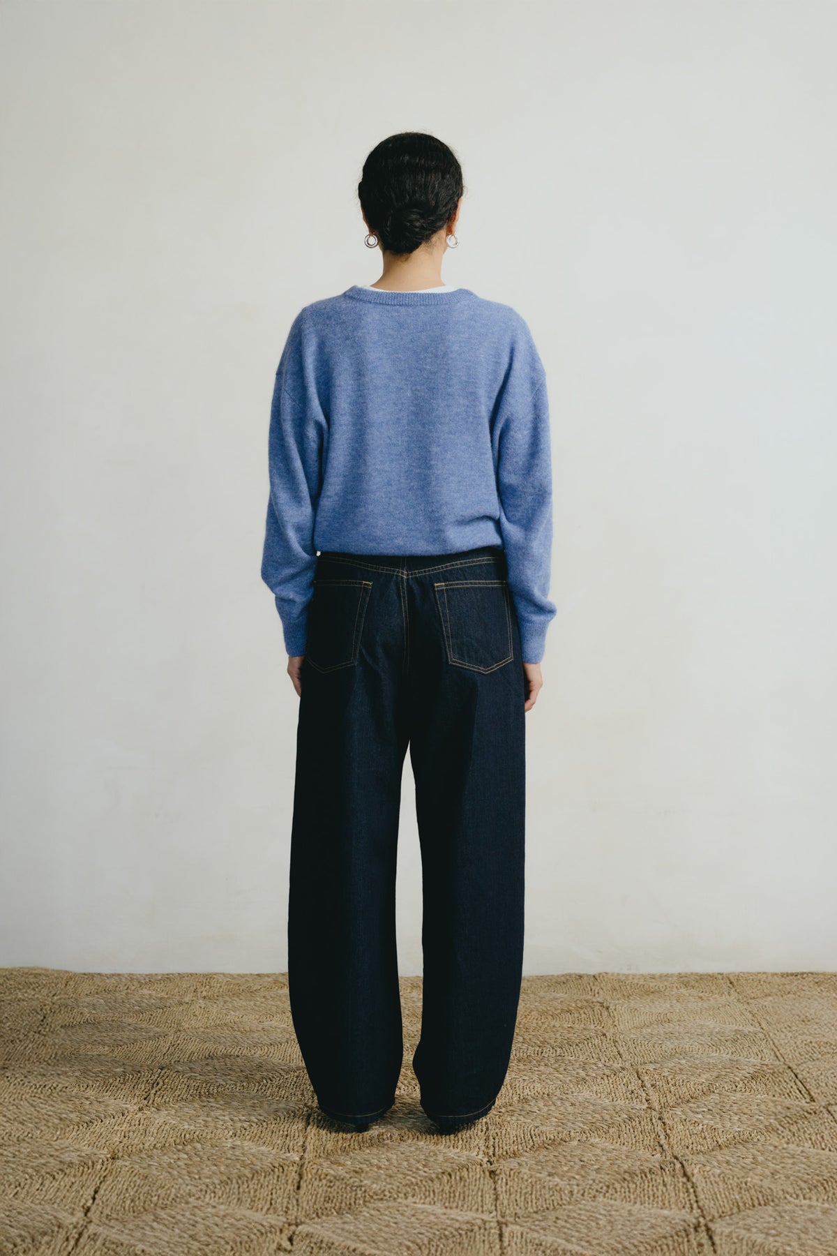A person standing with their back to the camera, wearing a heathered periwinkle blue long-sleeve knit sweater and dark wide-leg denim jeans, set against a plain off-white wall and a textured beige floor.