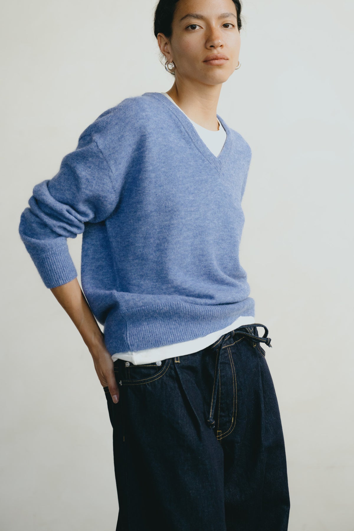 A model wearing a light blue heathered v-neck knit sweater over a white t-shirt and dark blue jeans, photographed against a plain off-white studio background.