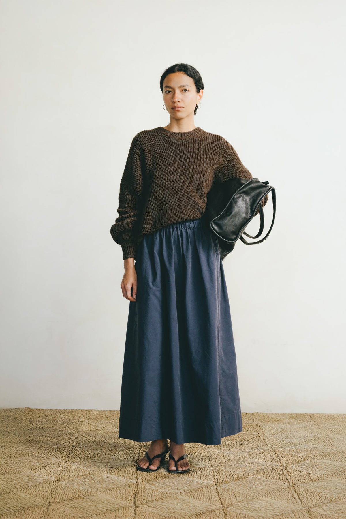 A woman stands against a plain off-white wall on a woven straw floor, wearing a dark brown ribbed knit sweater and a long navy blue skirt. She holds a black shoulder bag and wears black sandals.