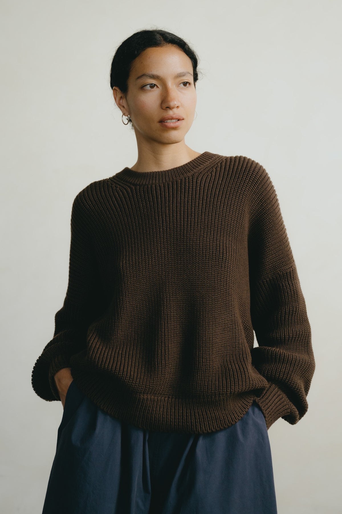 A woman wearing a dark brown ribbed knit crewneck sweater and navy blue pants, standing against a plain light gray background.