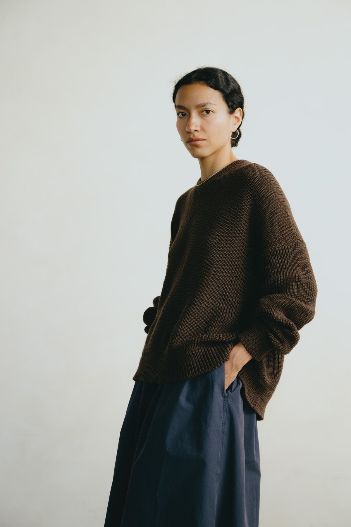 A model wearing an oversized dark brown ribbed knit sweater and a dark navy skirt, standing against a plain off-white background.
