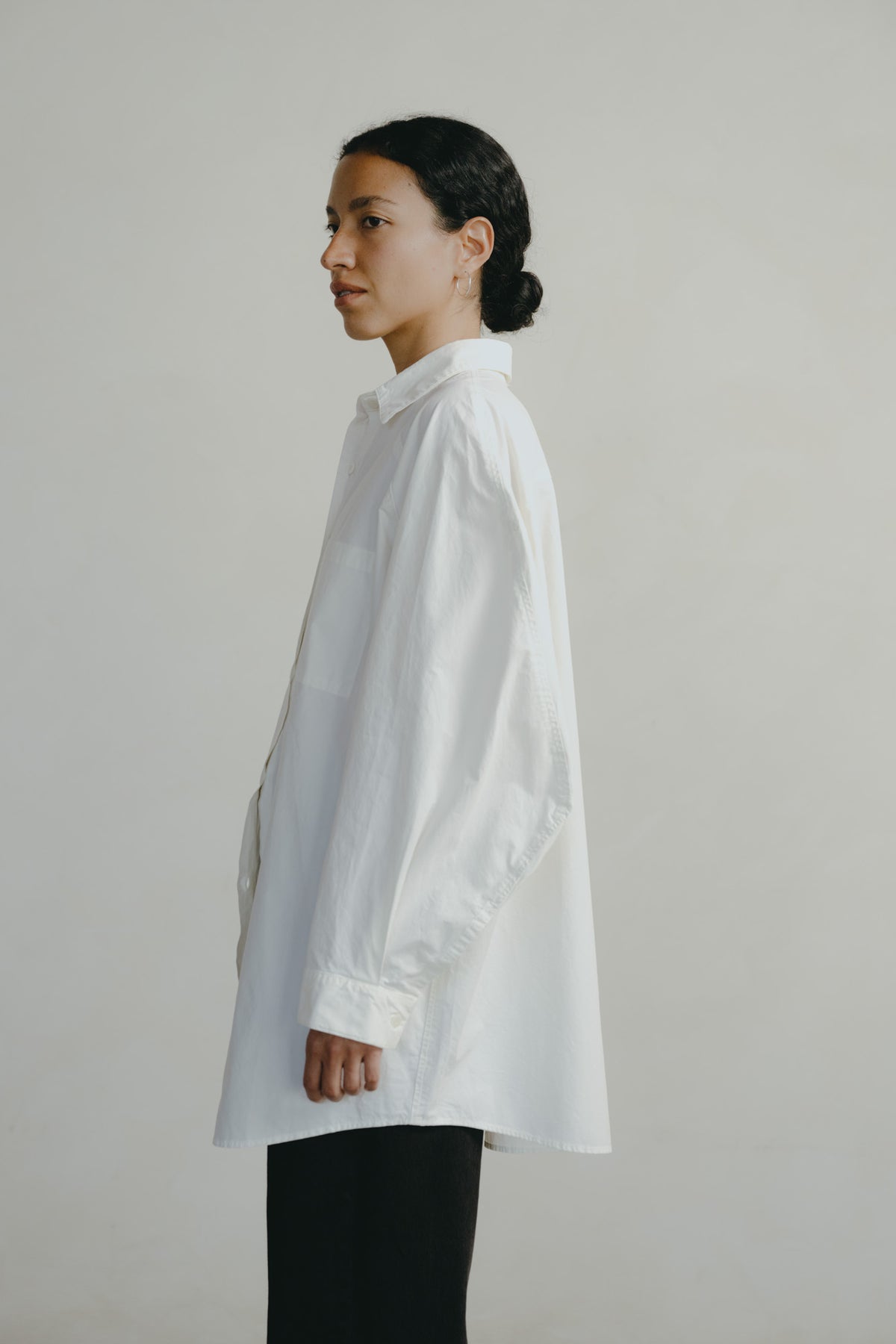 A woman in profile wearing an oversized white long-sleeve button-up shirt and black pants, standing against a plain light gray studio background.