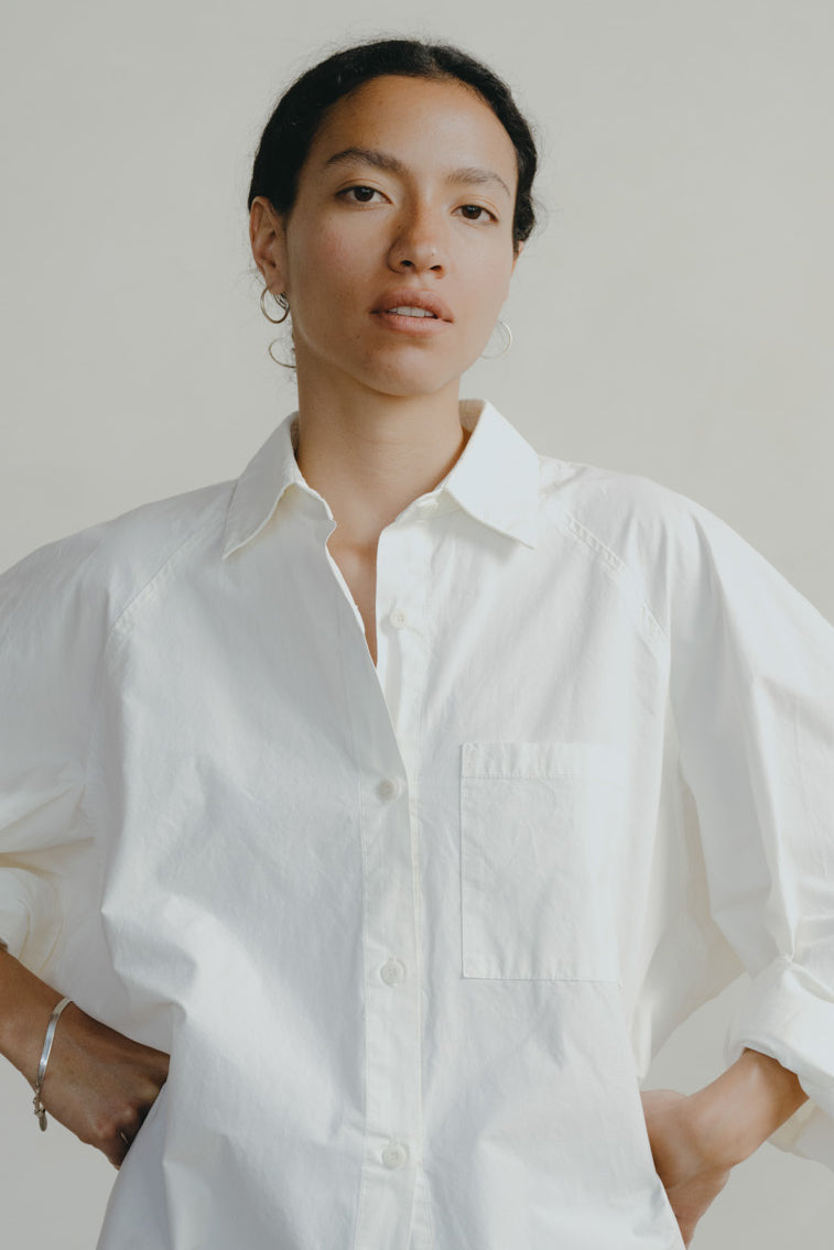 A woman wearing a white long-sleeve button-up shirt with a chest pocket, photographed against a plain light beige studio background.