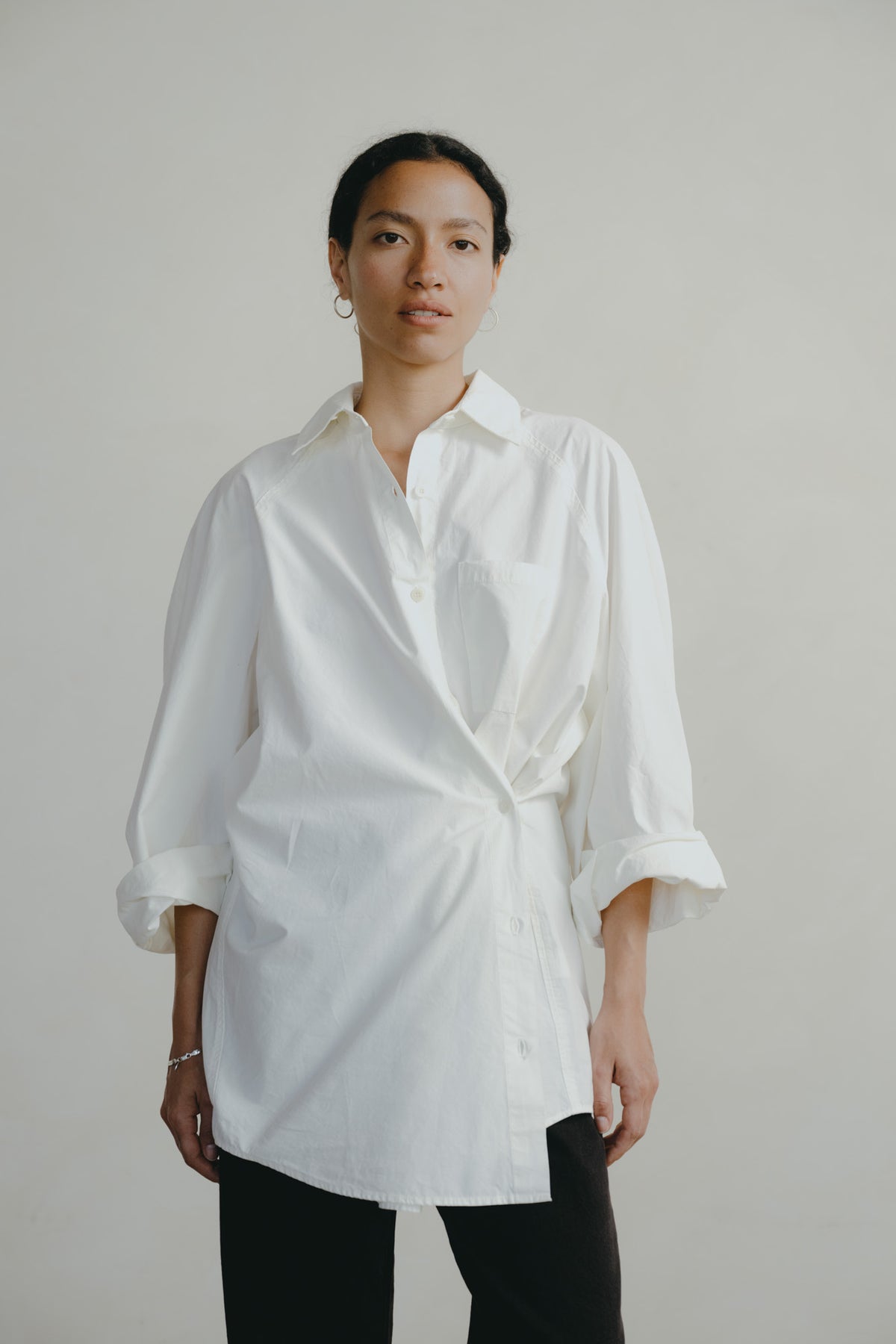 A woman modeling a white oversized button-down shirt with rolled three-quarter sleeves and a self-tie waist, paired with black pants, against a plain light gray studio background.