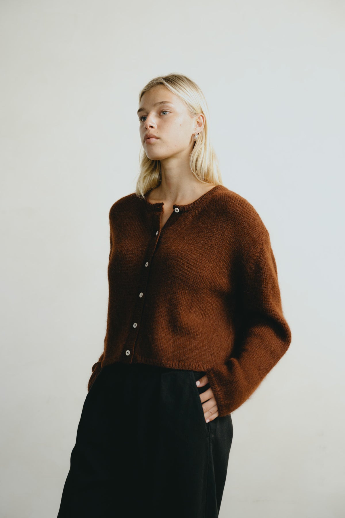 A blonde woman wearing a brown fuzzy knit button-up cardigan and black trousers, standing against a plain light gray background.