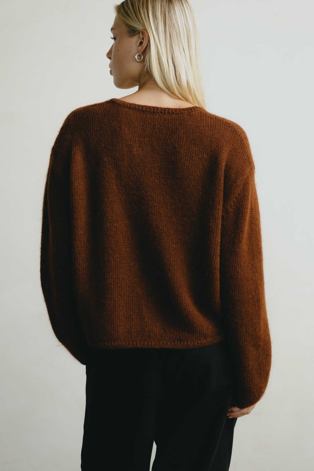 A woman shown from the back wearing a brown knit sweater and black pants against a plain light background.
