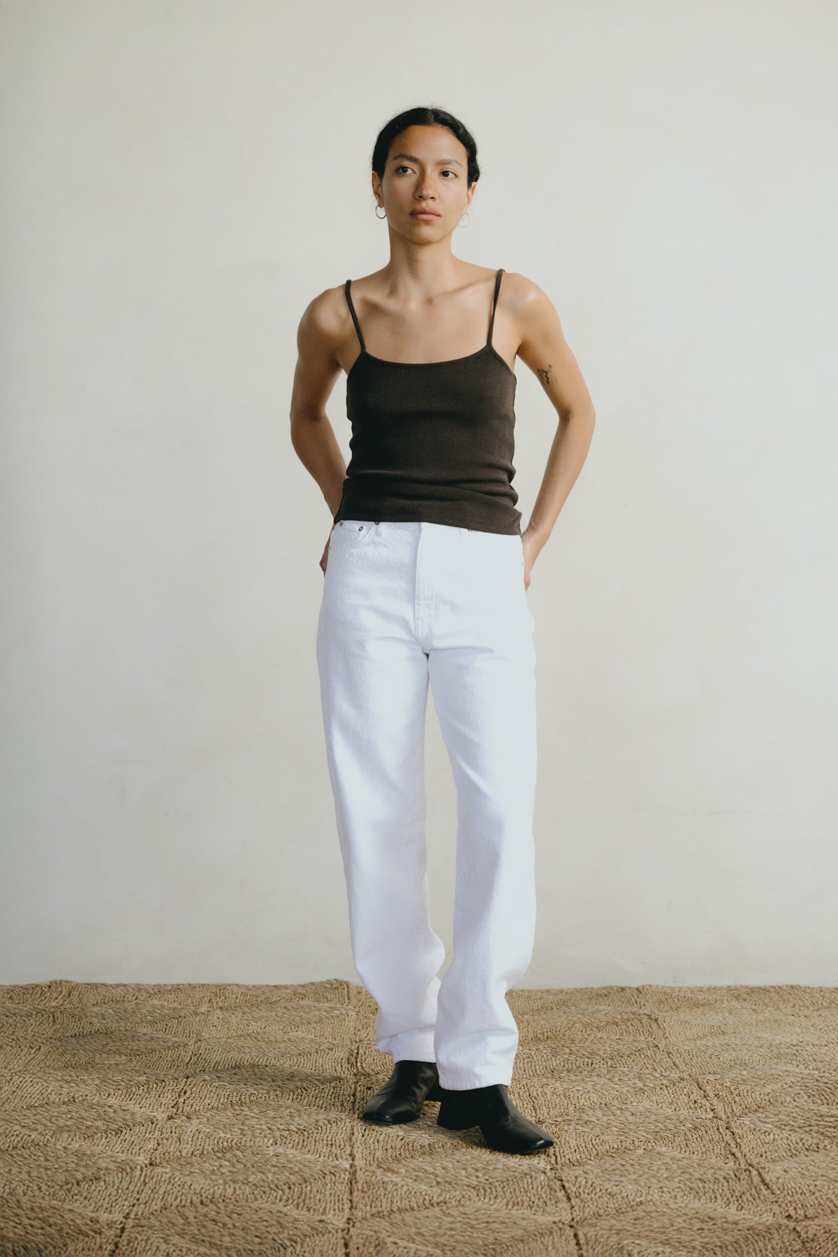A model stands in a studio wearing a dark brown ribbed camisole, white wide-leg trousers, and black shoes, set against a neutral off-white wall and a textured tan floor.
