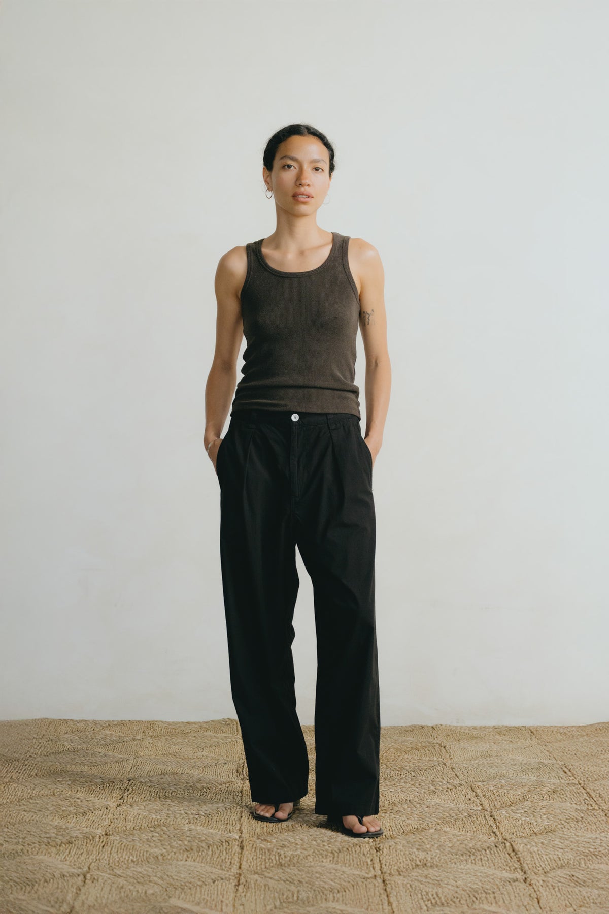 A model stands in a minimalist studio setting wearing a dark brown ribbed tank top and loose black wide-leg trousers, paired with black sandals on a textured beige floor against a light gray wall.