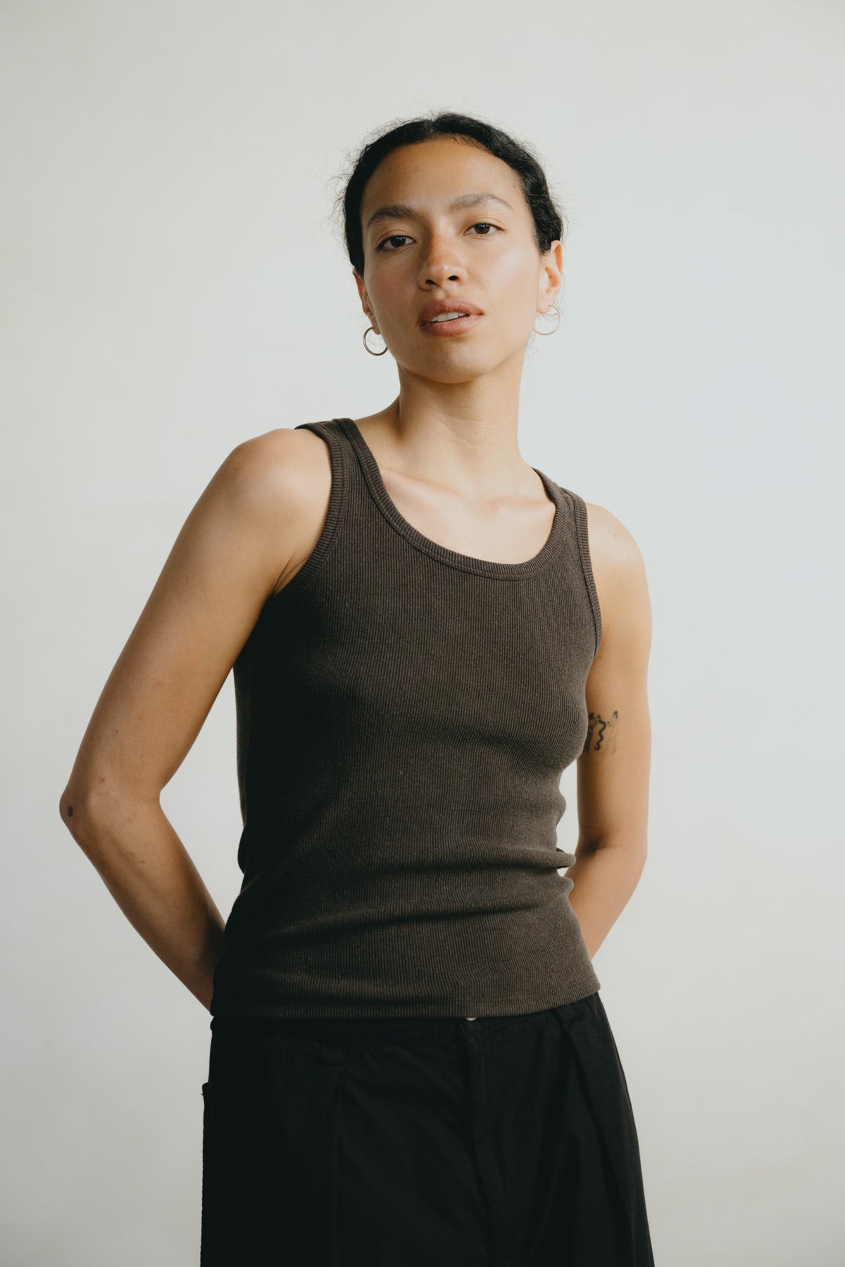 A woman wearing a dark brown ribbed tank top and black pants, standing against a plain light gray background.