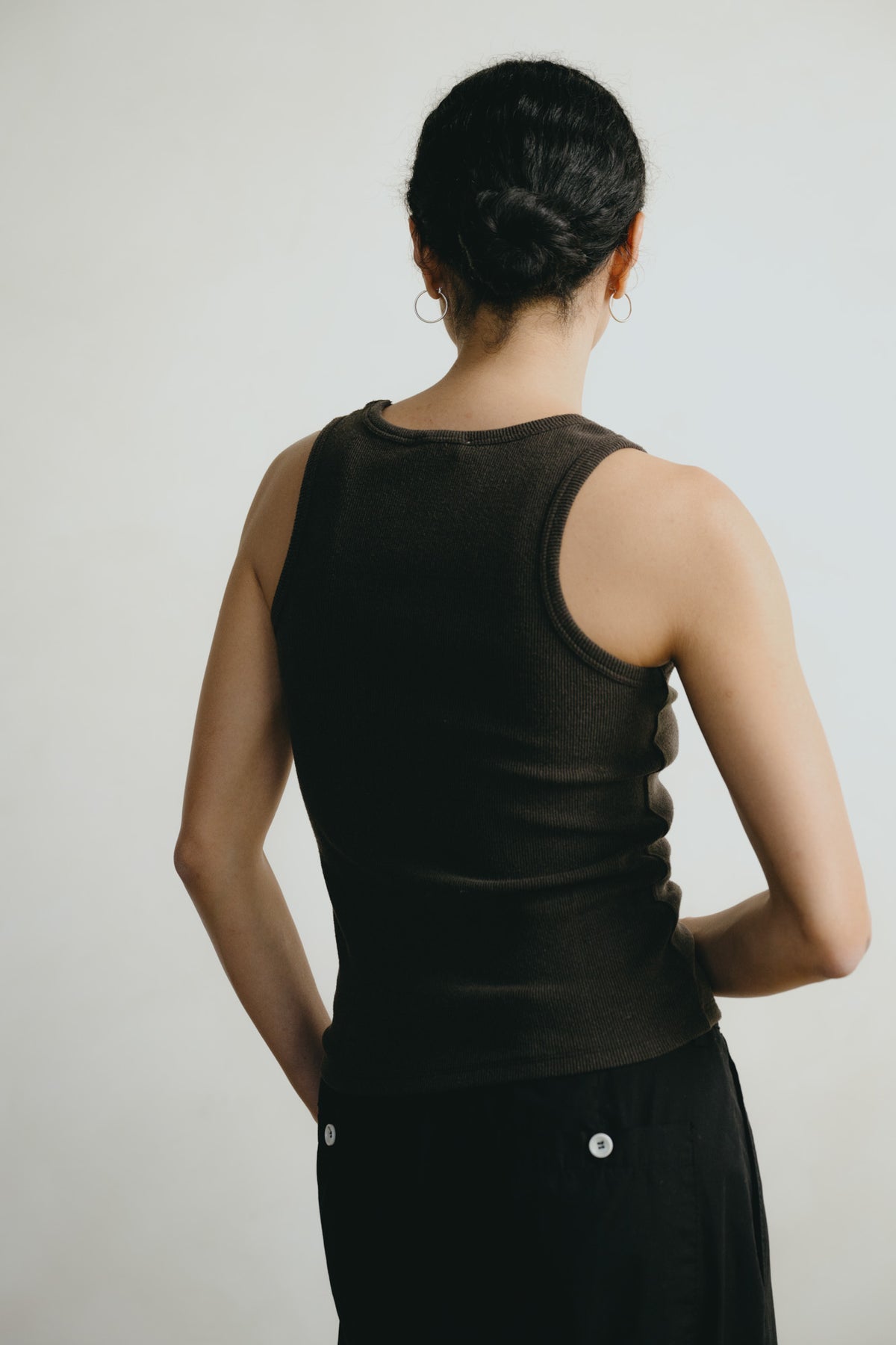 A woman shown from behind wearing a dark brown ribbed knit tank top and black pants against a plain light gray background.