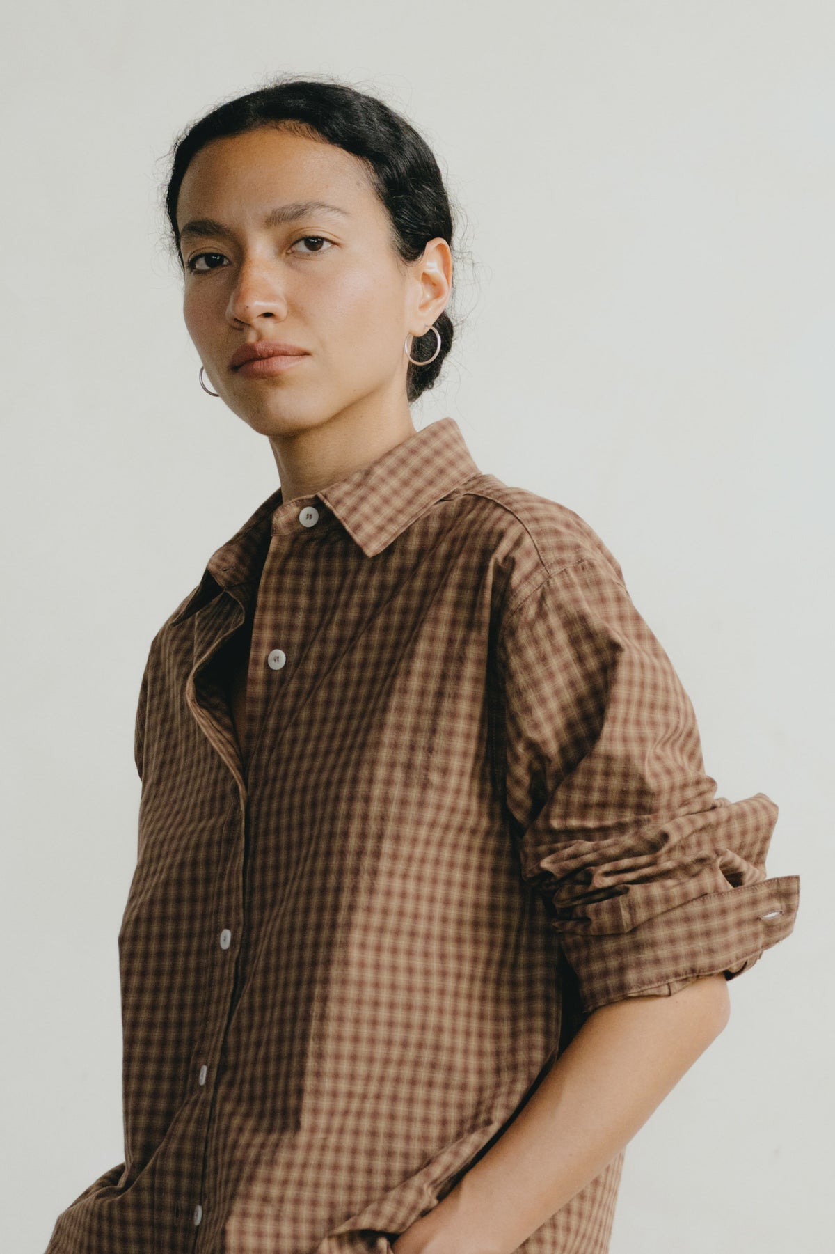 A model wearing a brown and beige checkered button-down shirt with rolled-up sleeves, photographed against a plain light gray background.