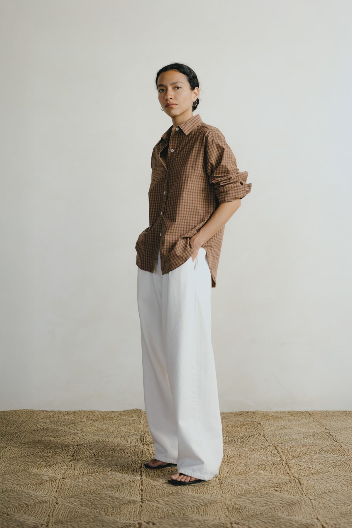 A model stands in a studio wearing a brown and beige checkered button-down shirt with rolled-up sleeves and white wide-leg trousers, standing on a textured straw-colored floor against a plain off-white wall.