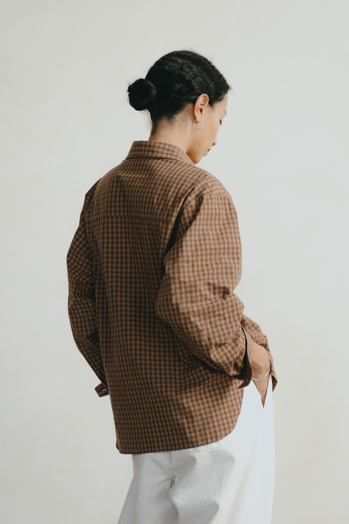 A woman shown from the back wearing a brown and beige gingham button-up shirt and white pants, standing against a plain light gray background.
